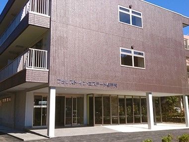 施設が建っている。左下側には玄関があり、その上側には屋根がある。外壁の左下側には施設名が書かれている。