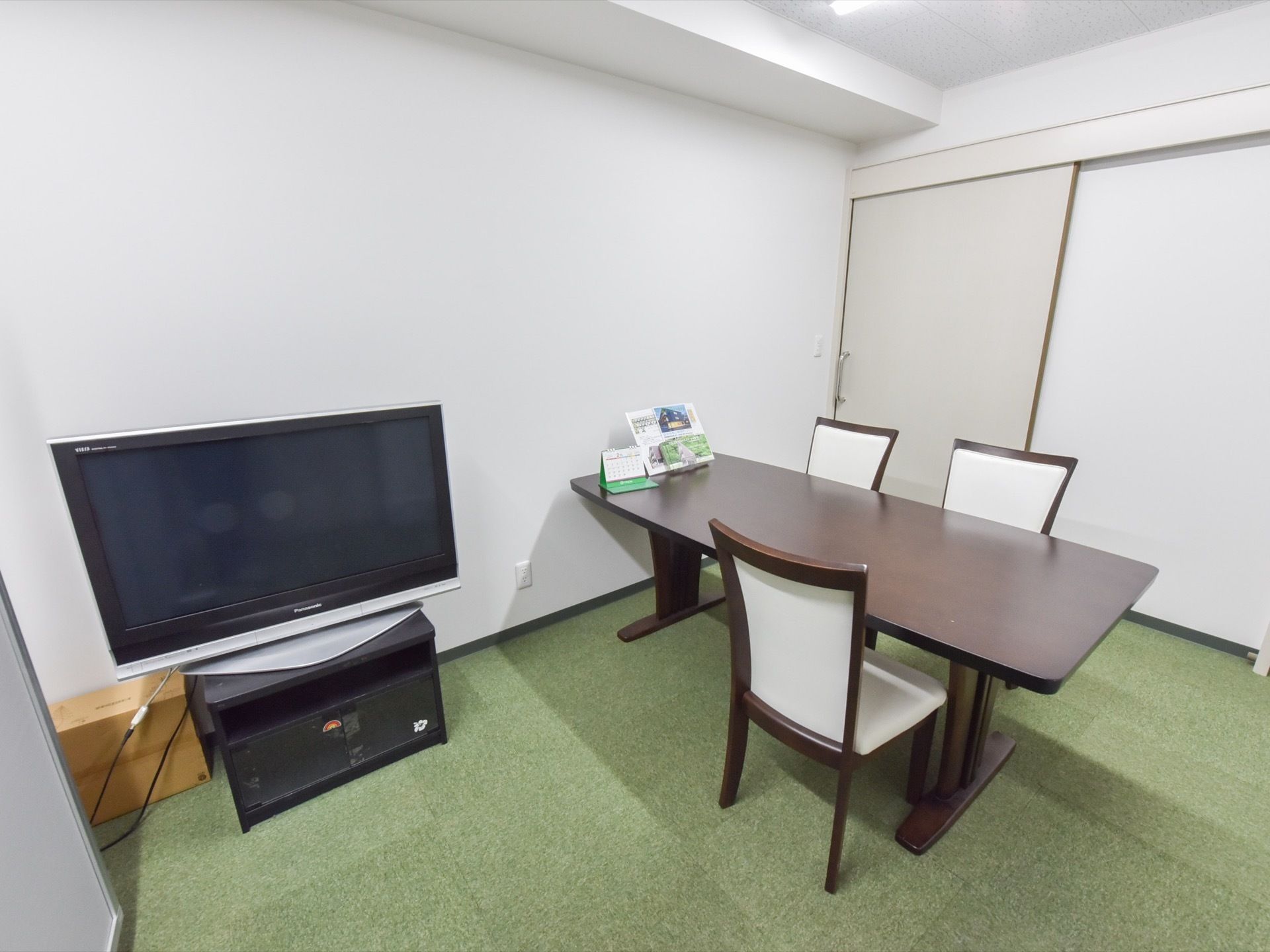 白い壁に緑のカーペットの空間の中には、テレビ、テーブルに椅子が３脚設置されている。テーブルの奥には引き戸が付いている。
