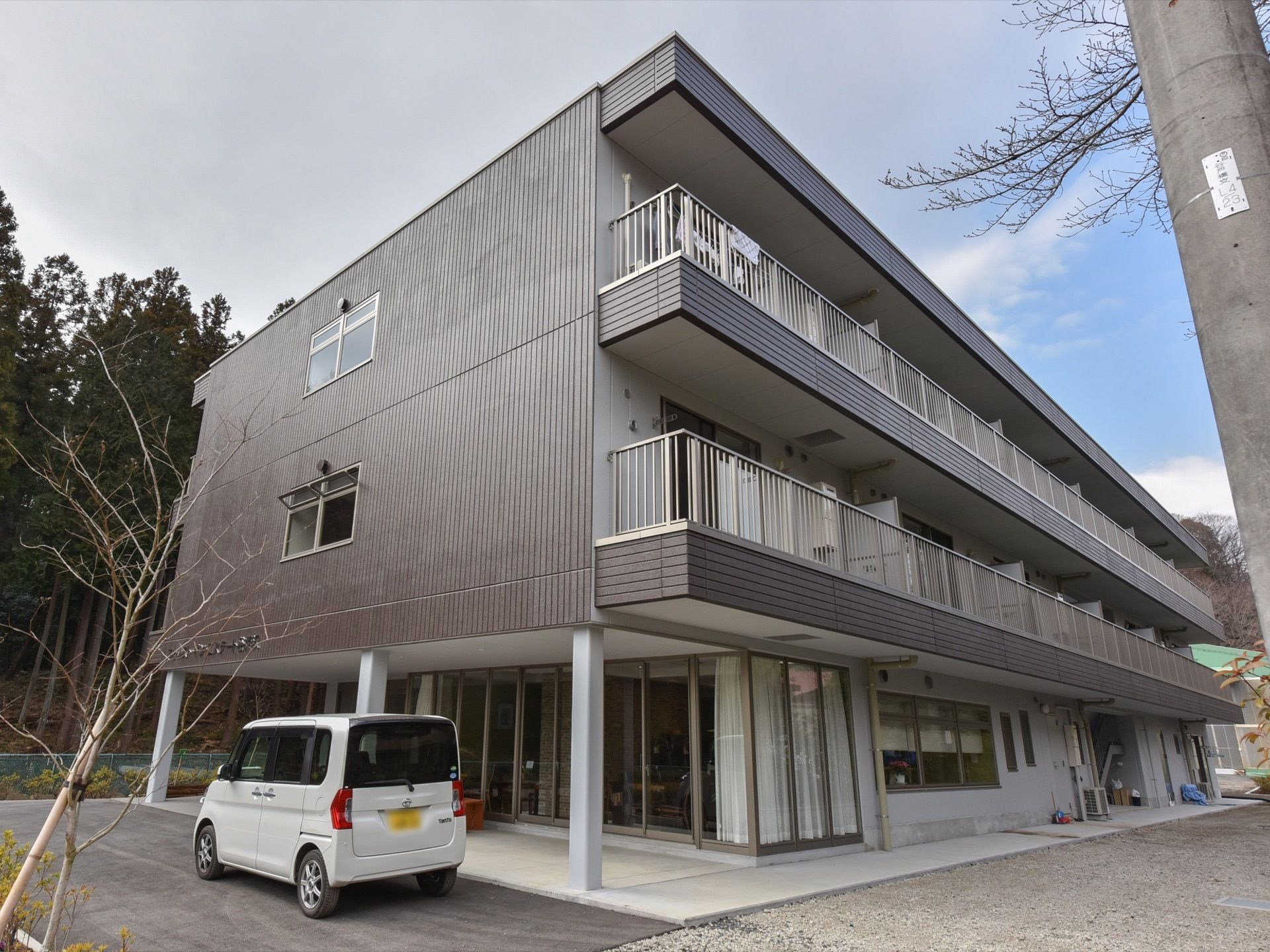 濃いブラウンの壁が特徴的な施設は３階建てである。各居室には、屋根の付いたベランダがついている。周辺は自然が溢れている。