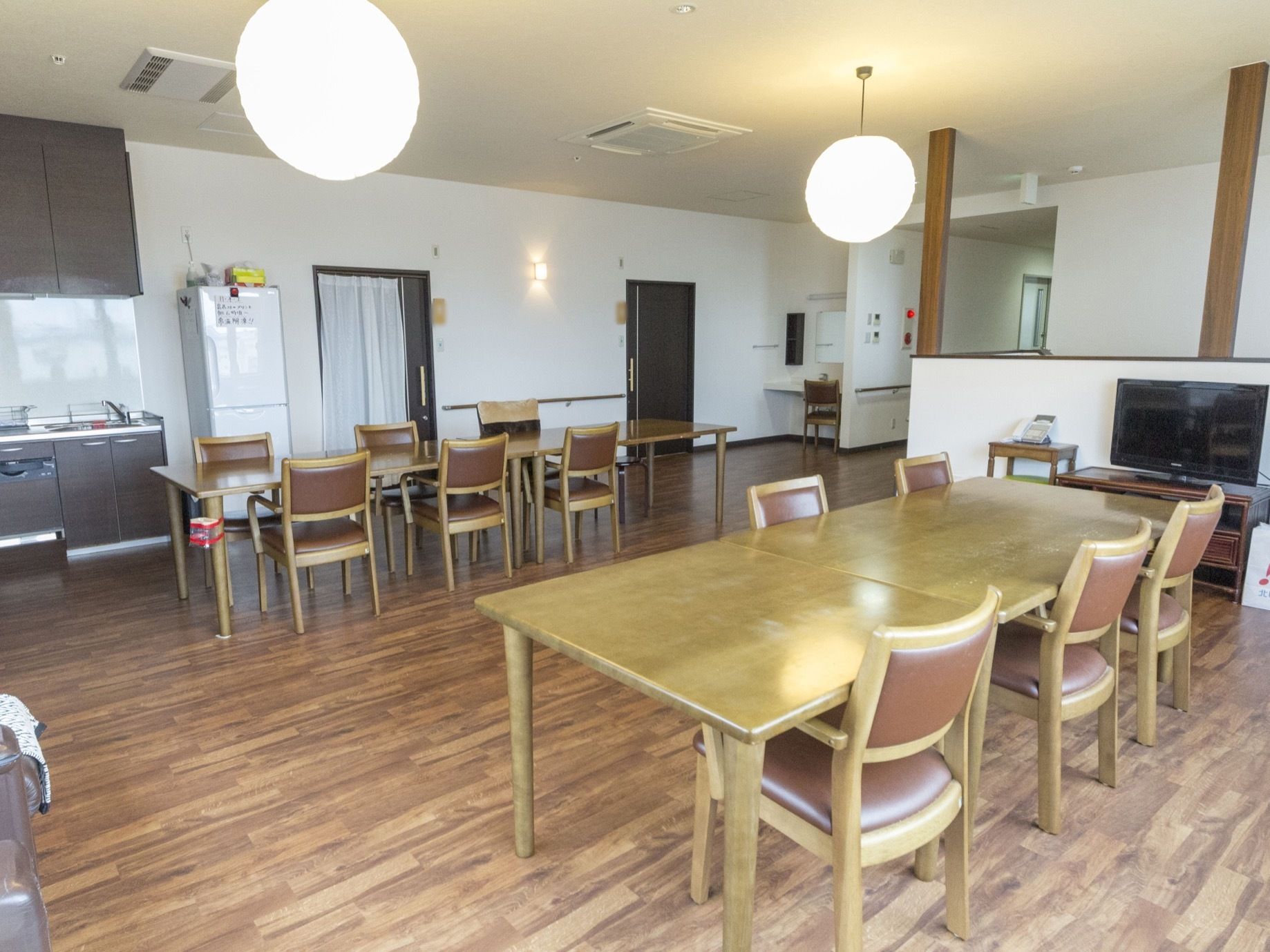 広々とした食堂には複数のテーブルセットが置かれている。一角にはキッチンがあり、隣には冷蔵庫が置かれている。