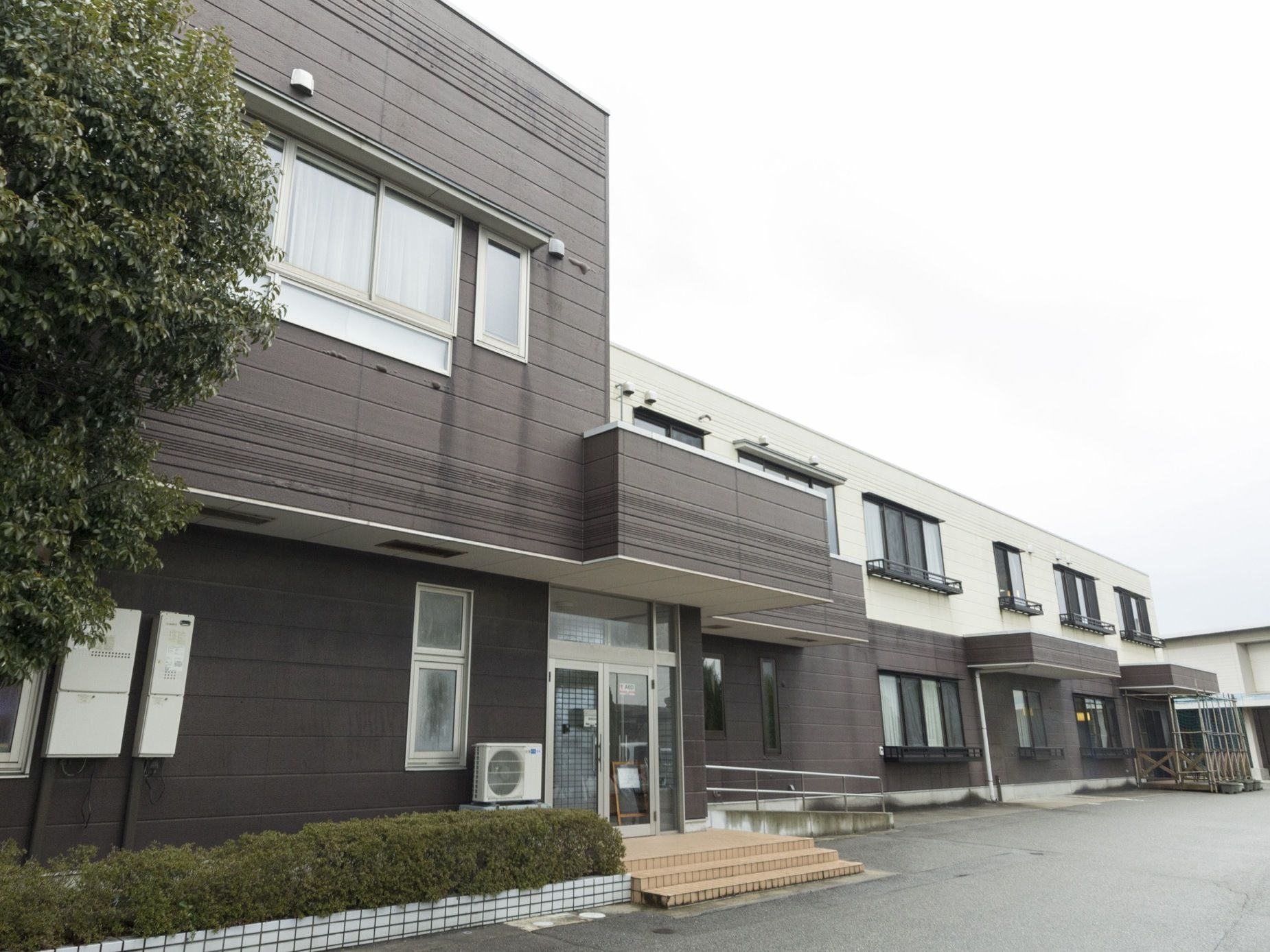 施設は落ち着いた色合いの2階建ての建物。入口前にはひさしがついている。施設の前は舗装されている。一角には植栽がある。