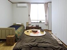 奥に窓があり、その手前に丸い天板のこたつ、左にベッドが置かれた居室