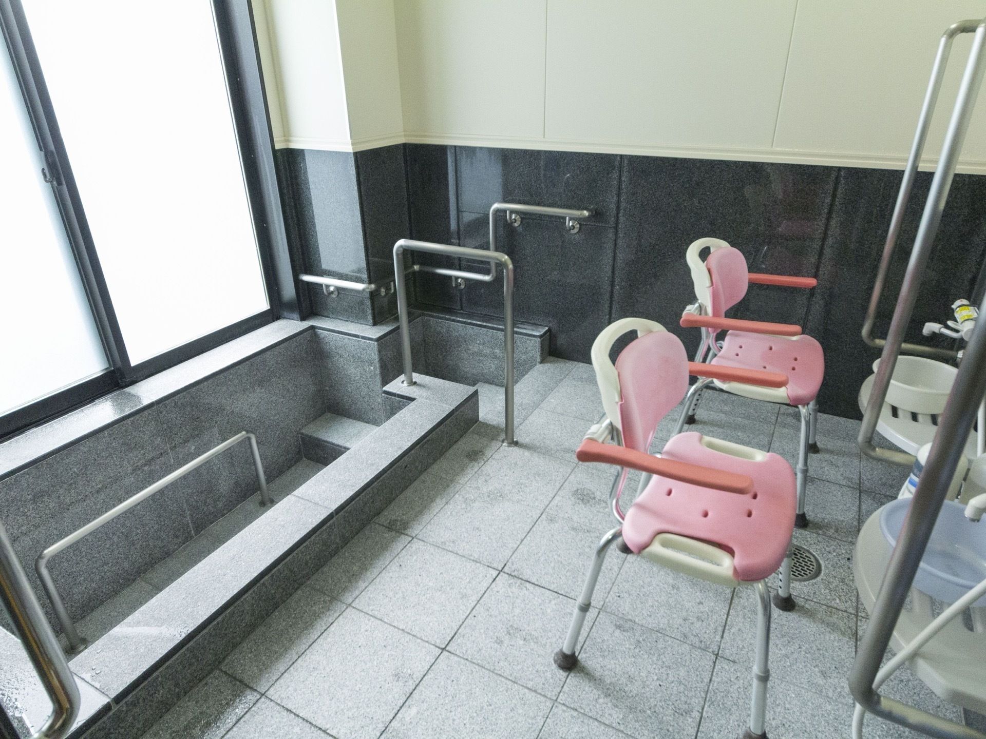 左に窓、その下に石造り風の浴槽があり、右側にピンクの浴用椅子が2つ置かれた浴室