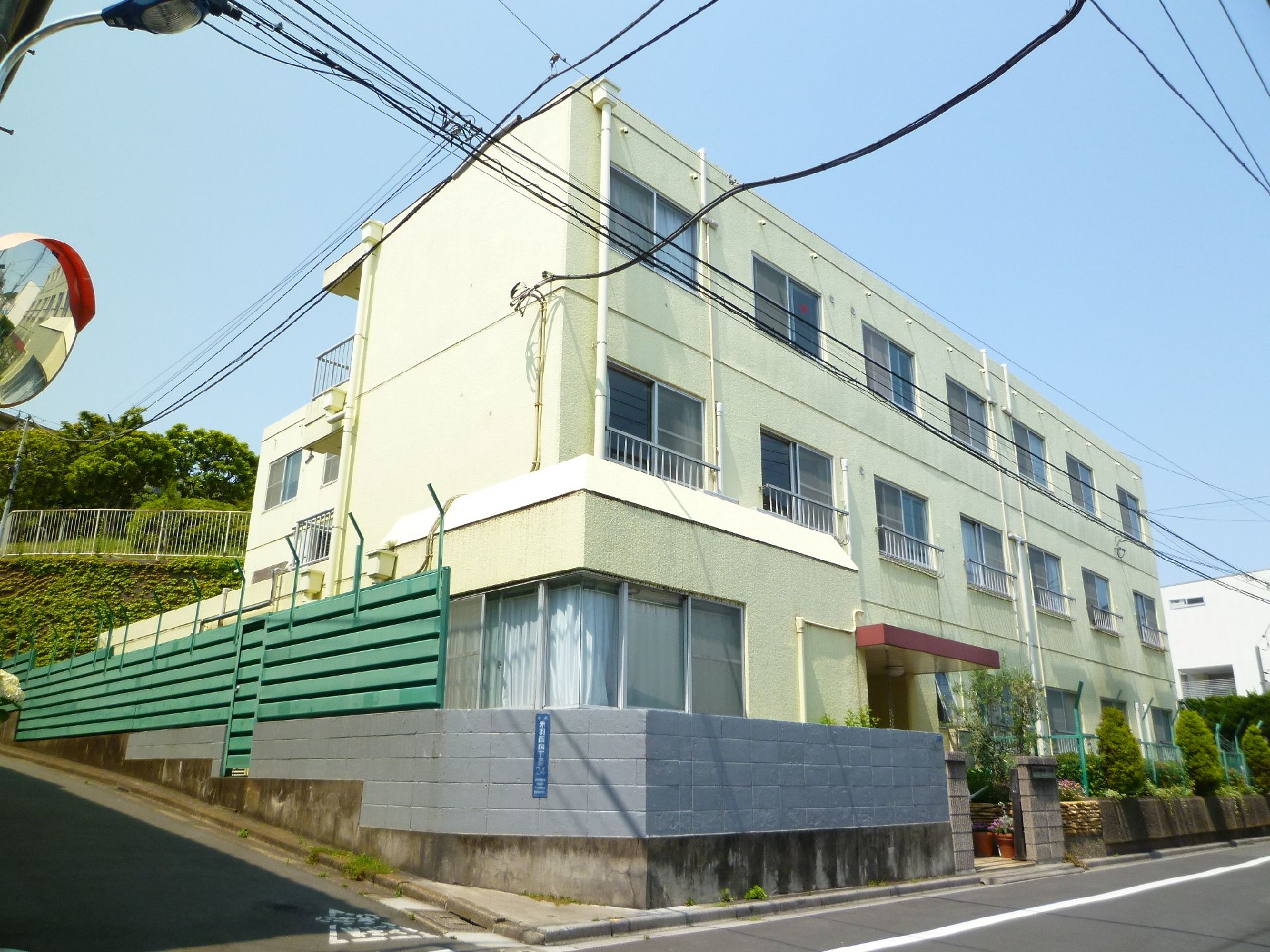 左に登る坂道と右に伸びる道路の角地に立つ3階建ての白っぽい建物