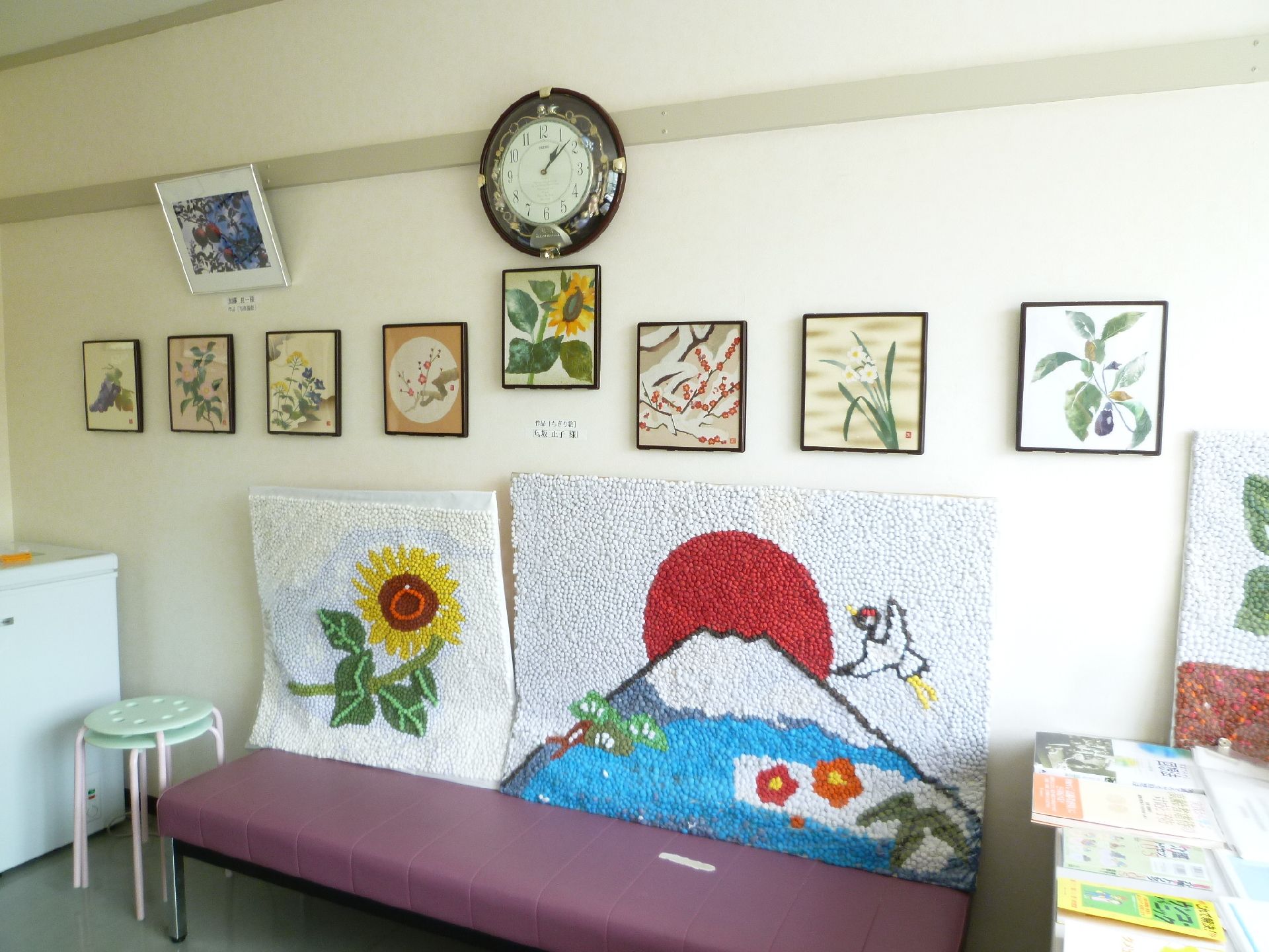 白い壁面にちぎり絵の作品が飾られ、その手前の紫のベンチの脇にも作品のあるスペース