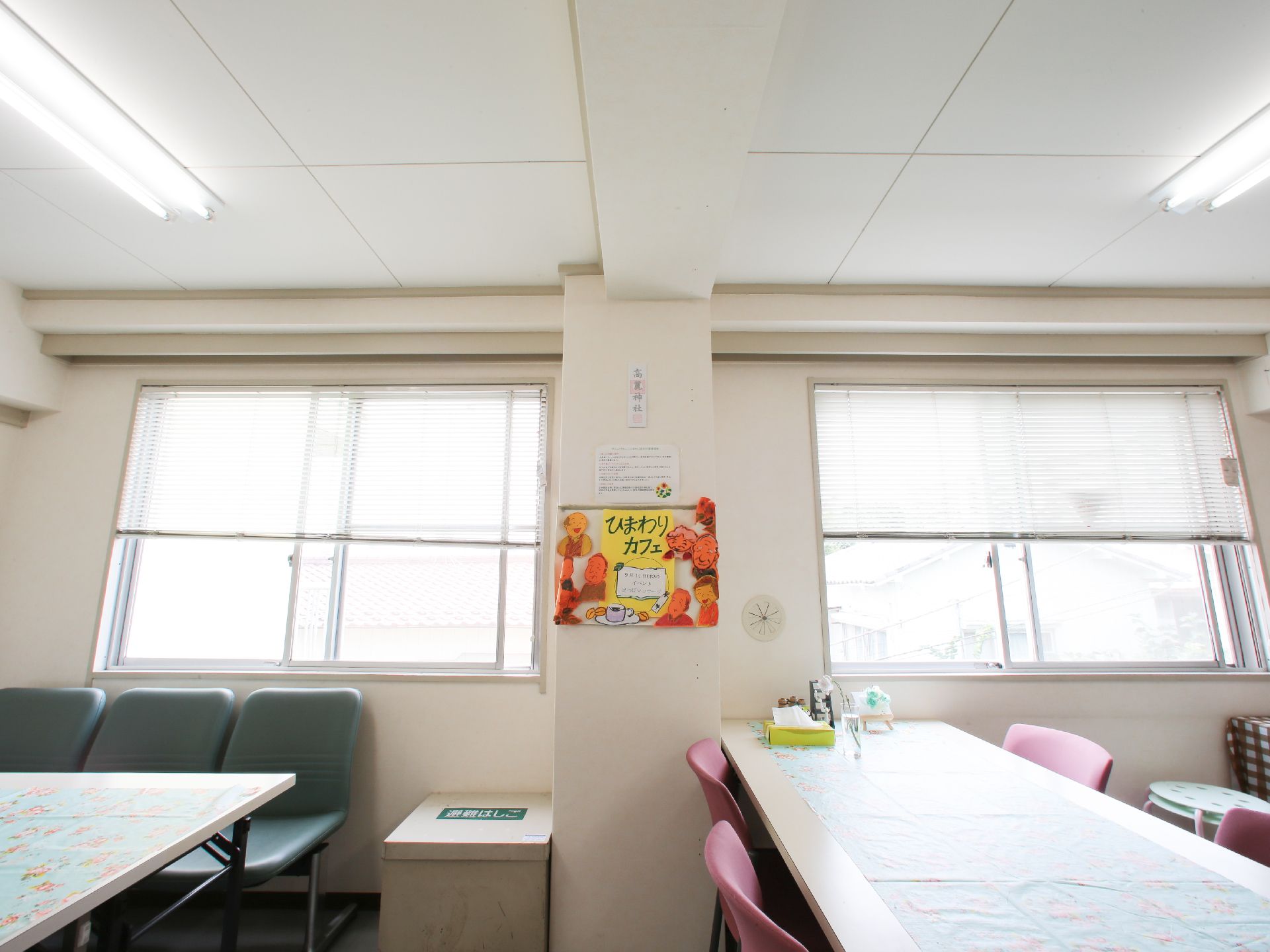 窓にはブラインドが取り付けてある。白い壁にはひまわりカフェというポスターが貼られている。テーブルとイスが設置されている。