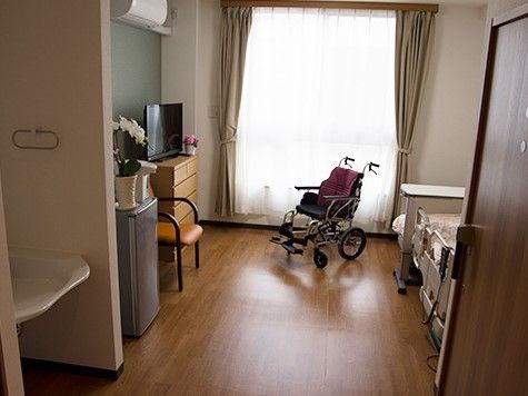 正面に窓があり、その前に車椅子が置かれ、右壁面にテーブル付きのベッドがある居室