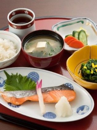 大根おろしと生姜を添えた焼シャケをメインに、ご飯、味噌汁、青菜のお浸し、人参ときゅうりの漬物が色鮮やかに並ぶ和食メニューである。