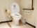 洋式トイレが設置されている。壁側にブラウンの手すりがあり、便器の右側には可動式のものが取り付けられている。
