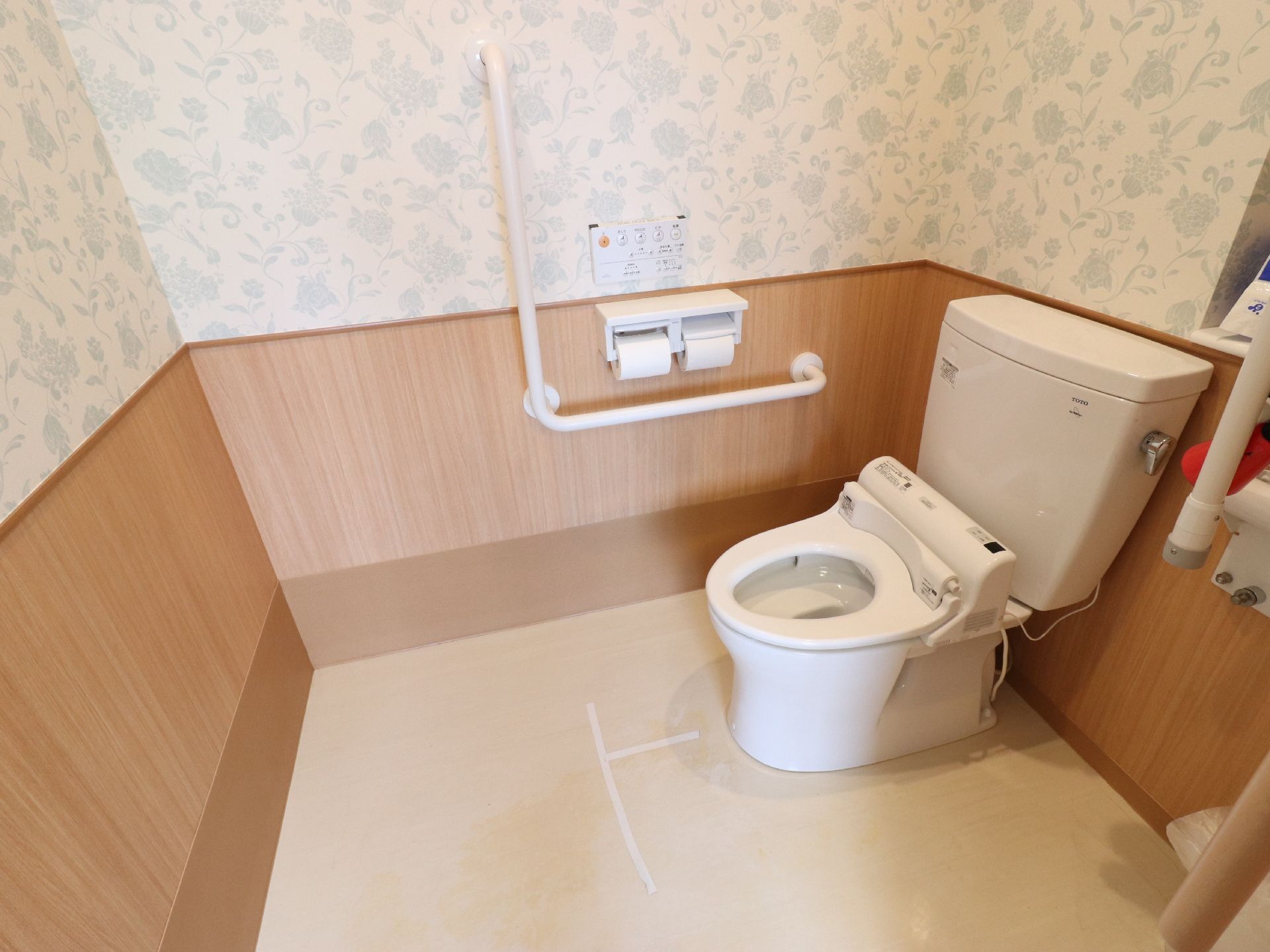 茶色の腰板に、ブルーの花柄のついた壁紙の個室である。温水洗浄器付き便座の洋式トイレで、壁に手すりが設置されている。