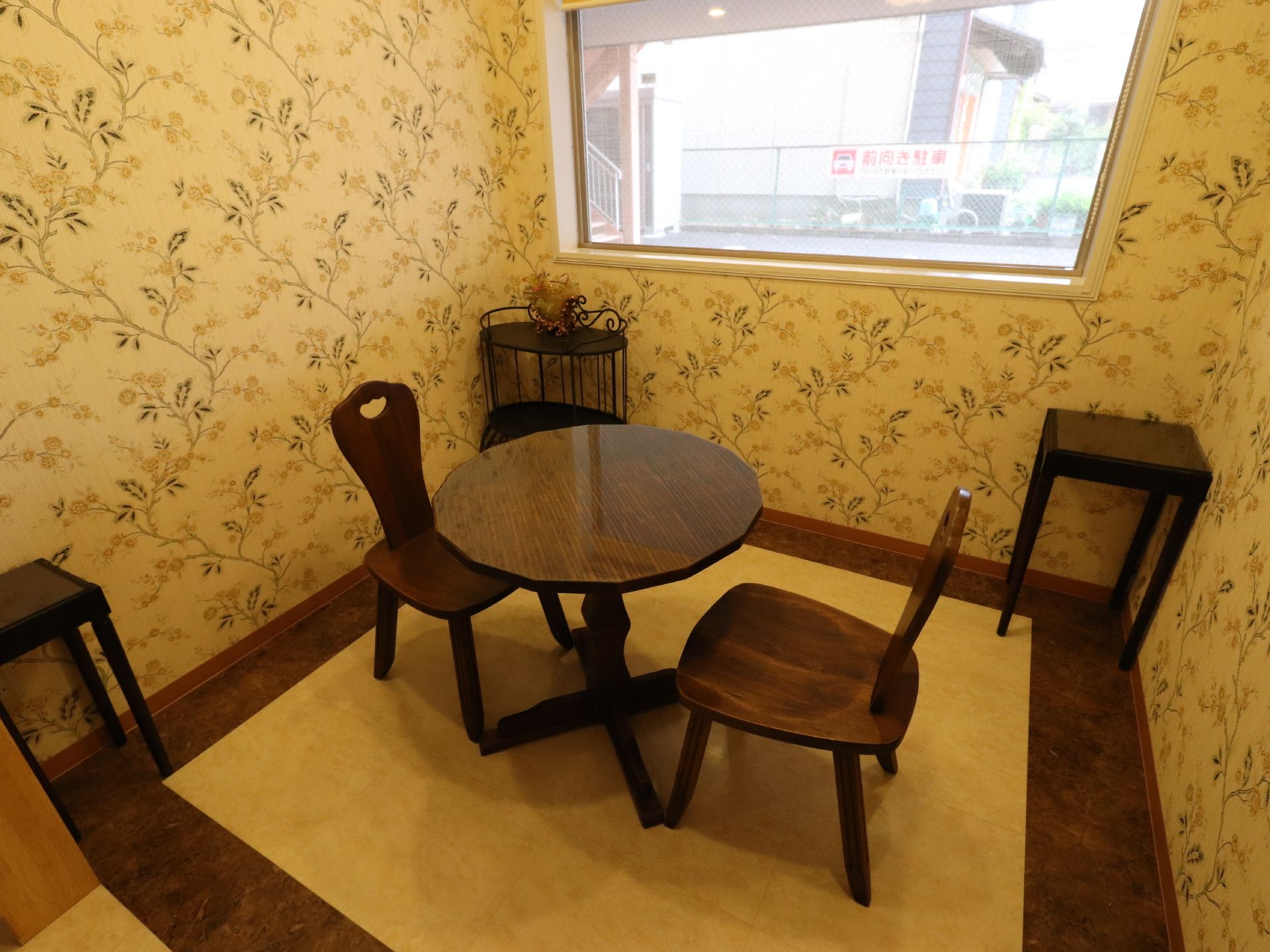 正方形の小さな部屋で、ツートンカラーの床に花の模様の壁紙である。木の椅子と丸テーブル、サイドテーブルなどが置かれている。