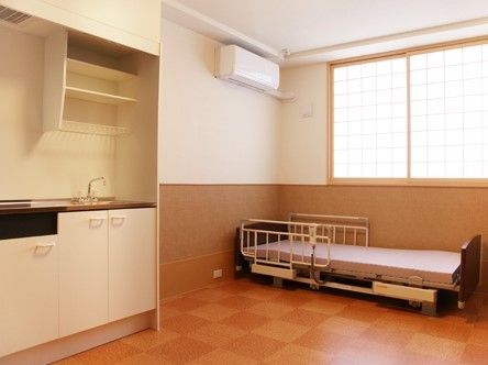 茶とベージュの大きな石畳模様の床、障子付きの窓で和を感じさせる部屋である。ミニキッチンとエアコン、電動リクライニングベッドがある。