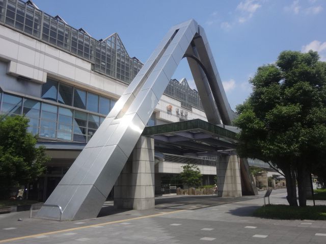 駅舎の屋根部分と駅の入り口は三角を取り入れたデザインである。丸い植え込みスペースが両脇にあり、前方にも大樹がある。