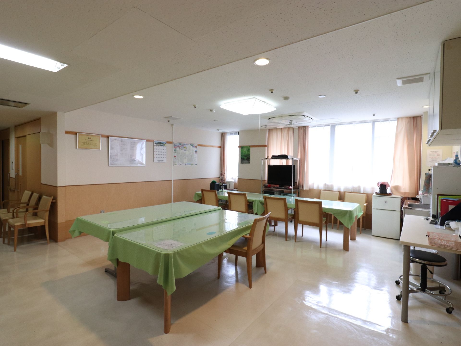 共同生活室の照明からはコードが垂れ下がっている。壁にはカレンダーなどが貼られている。テーブルも複数置かれている。