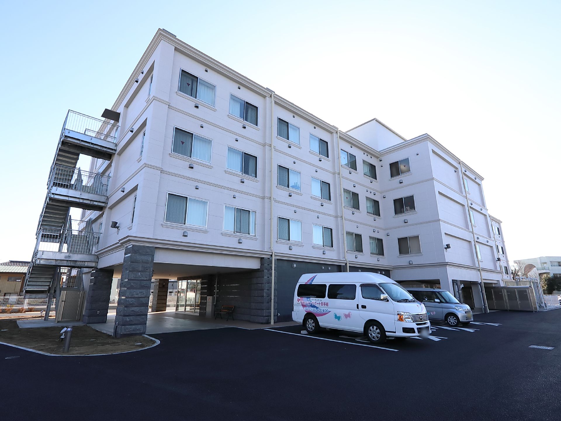 ４階建ての施設である。１階を掘り込んだ部分に玄関がある。施設名称が書かれた白色のワゴン車が玄関の近くに停まっている。