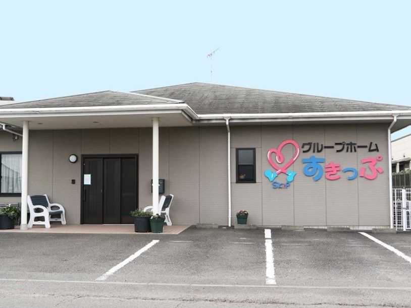 右端に施設名が入ったグレーの壁に、ライトグレーの寄棟屋根になった平屋の建物