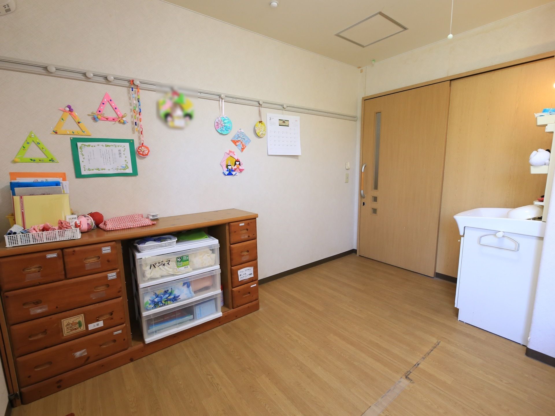 右奥に引き戸の入口扉があり、その前に白い洗面台がある。壁際に棚が置かれ、白い収納ボックスが収められている。