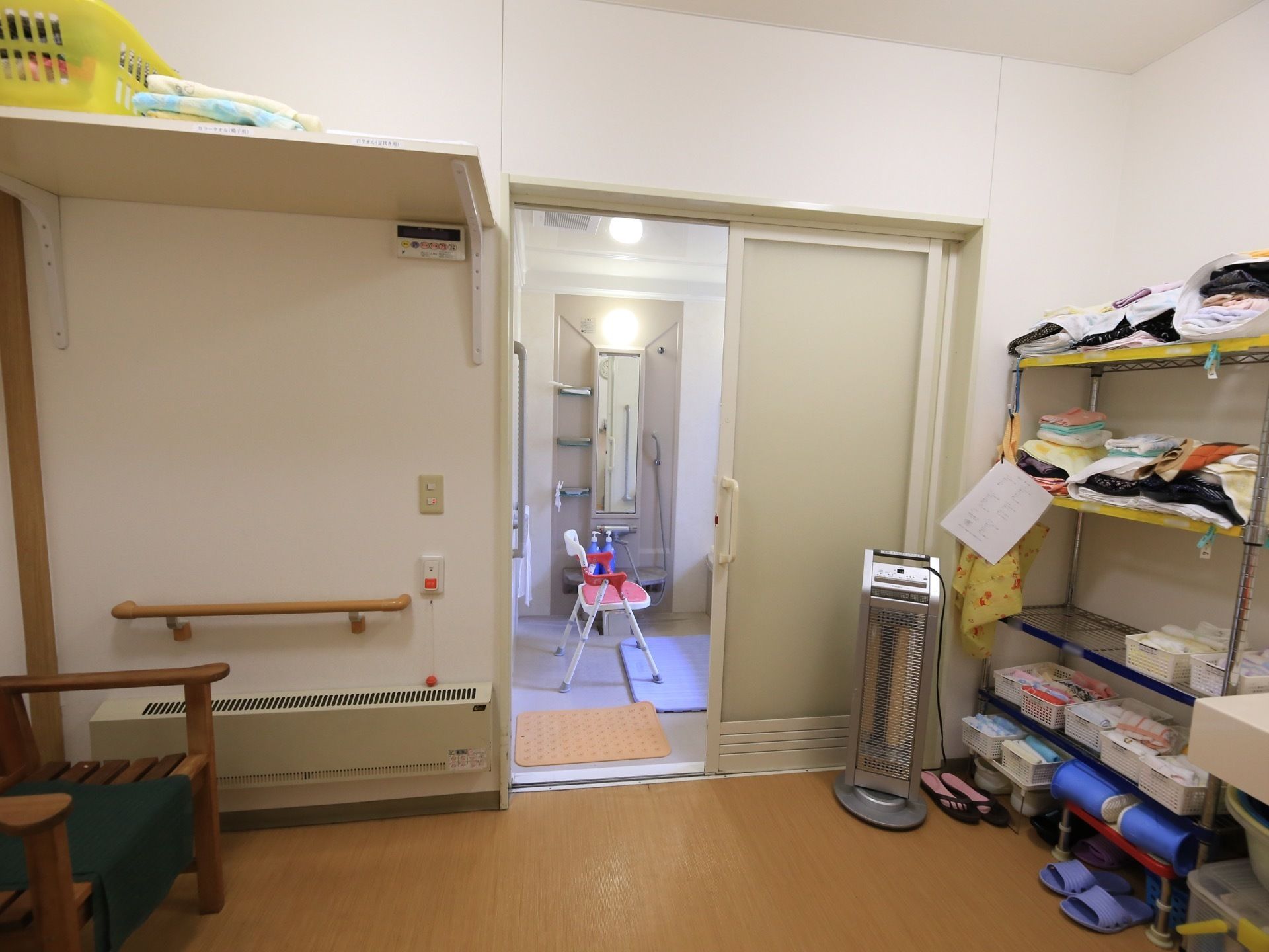 脱衣所には木製のベンチが１脚置かれている。床に近いところに暖房器具が取りつけられている。奥に浴室が続いている。