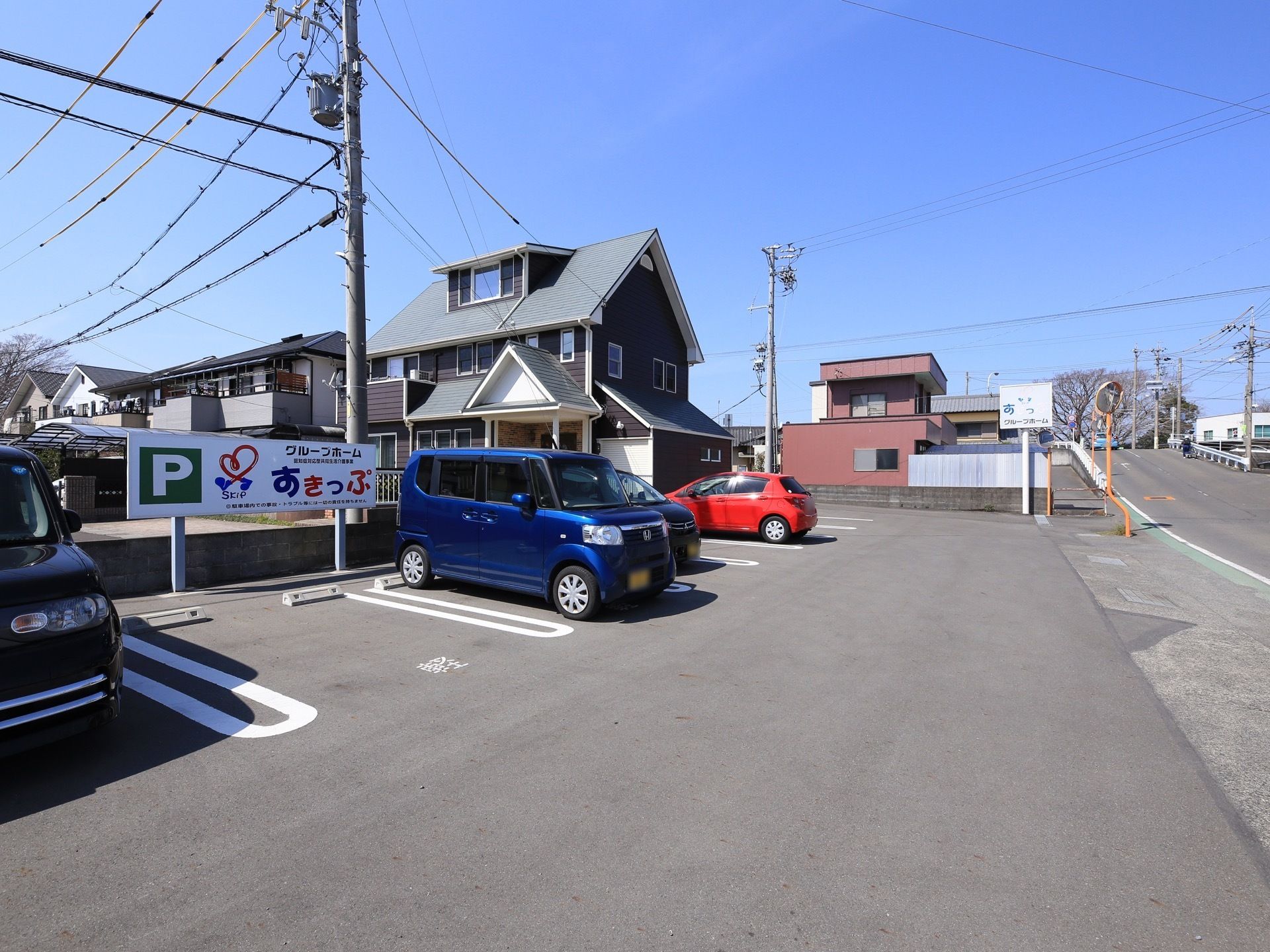 施設が保有する駐車場の写真である。通りに面して細長く敷地があり、青や赤、黒い自動車が停められている。