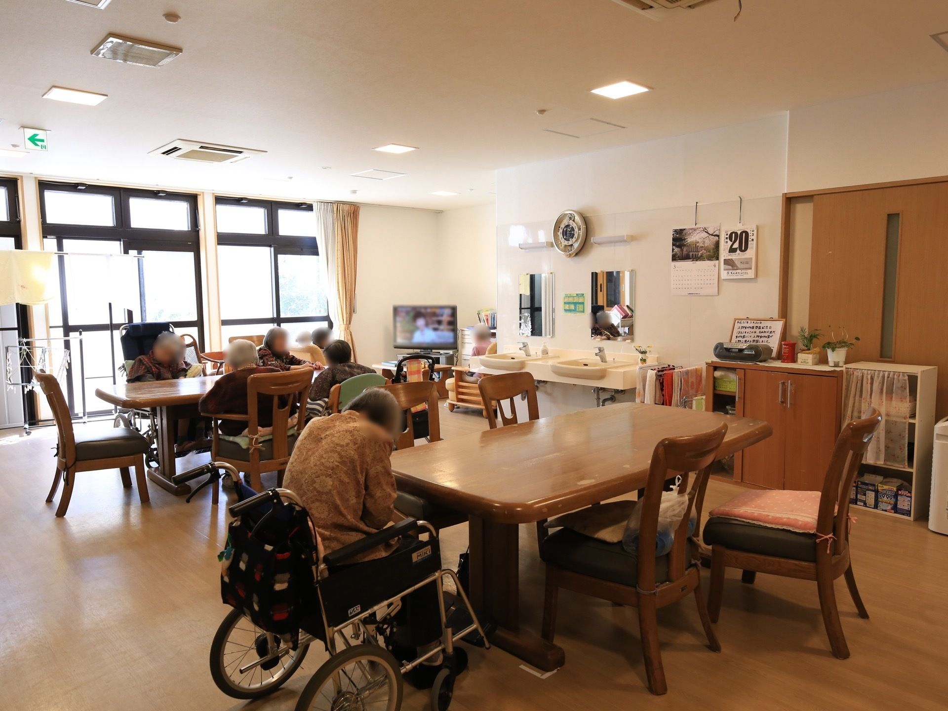 左奥にはたくさんの窓があり、外光が差し込んでいる。窓の横には大型液晶テレビが置かれている。木製のテーブルセットが並んでいる。
