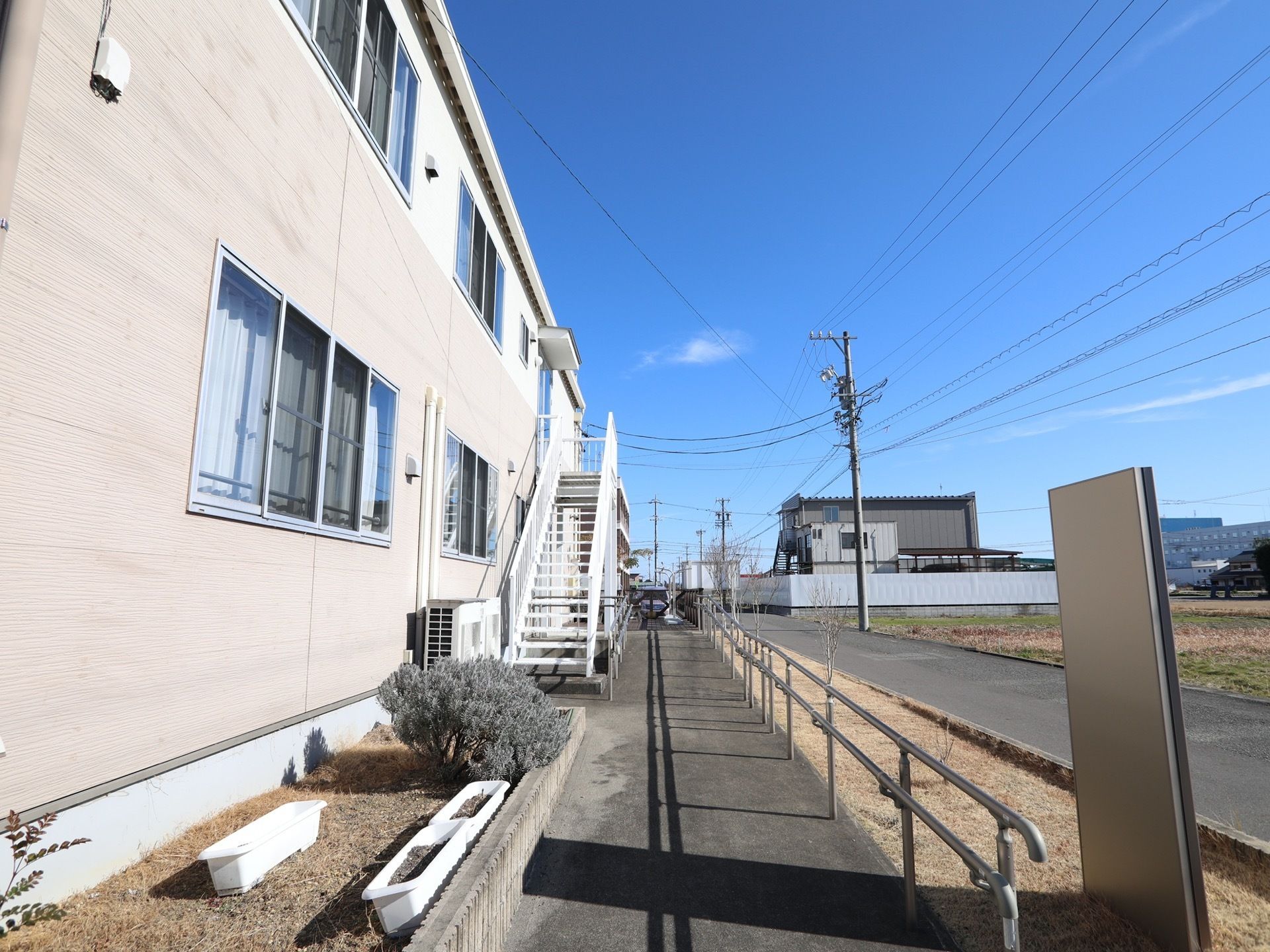施設前は広い田んぼが広がり、周囲にはあまり建物がない。歩道と車道の間には、柵と花木を植えるスペースがある。
