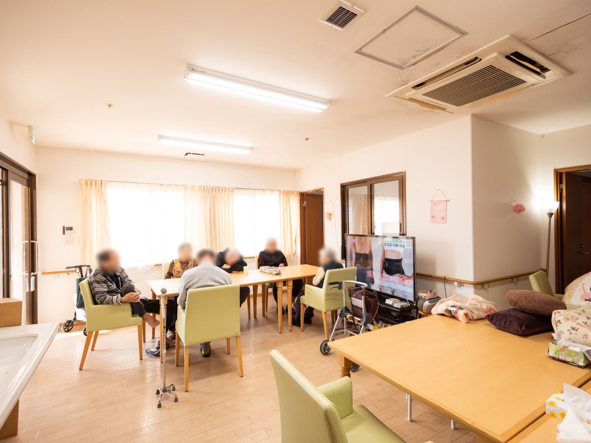 大きなダイニングテーブルを取り囲んで入居者様が複数人団らんしている。傍らに歩行器が置けるゆとりがある。天井に大型のエアコンが付いている。