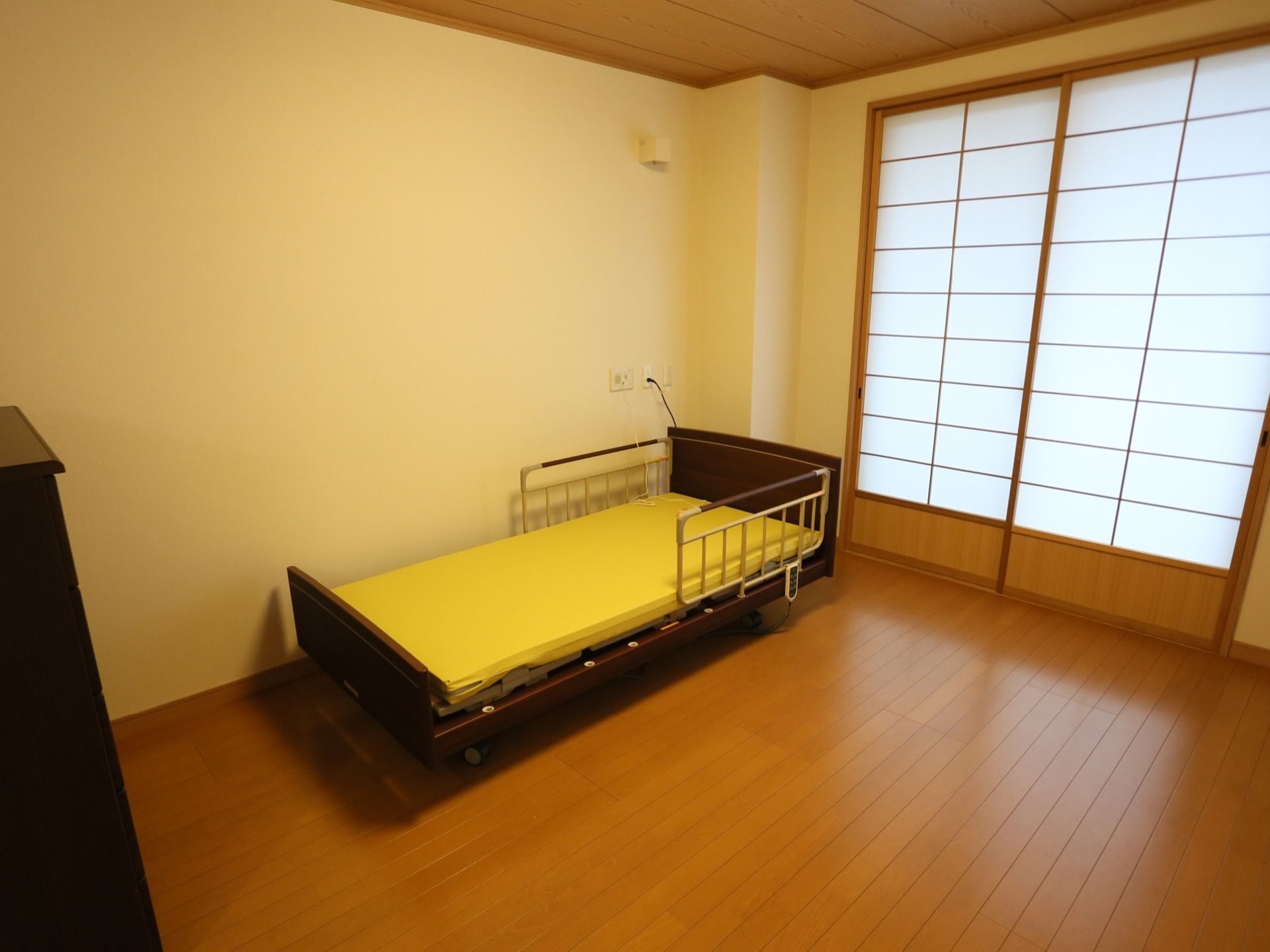 きれいなフローリングの居室に、介護ベッドや小ぶりの収納家具が置かれている。障子があり、天井は木質感がある。