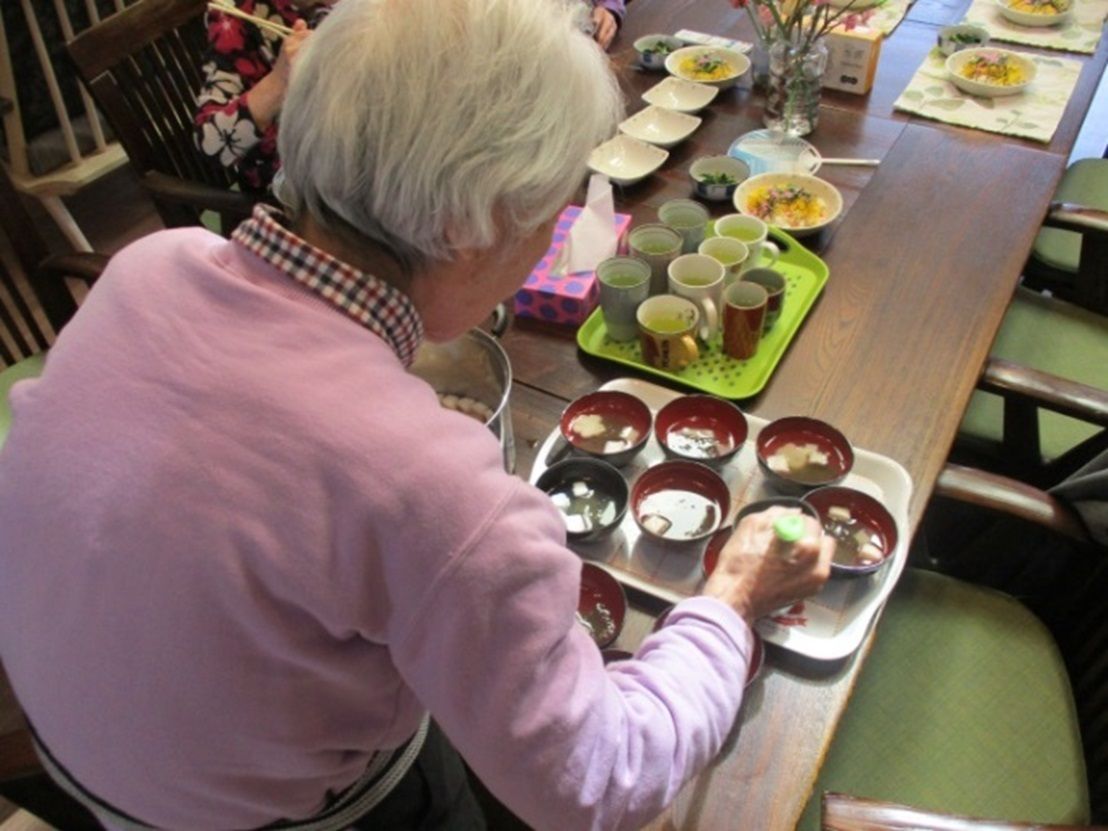 大きなテーブルの上で入居者様がみそ汁を椀につぎ分けており、次々トレーにのせている。ご飯はすでに各々の前に配られており、準備が進んでいる、