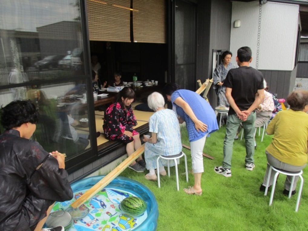 室内の引き戸は開け放たれて、縁側の雰囲気が作られている。入居者様とスタッフが交流を持ち、浴衣姿の人もいて夏祭りらしい賑いになっている。