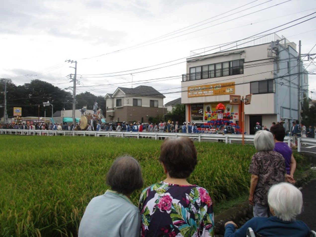 大行列が田んぼと住宅地の間の道路を進んでいる。人が乗った大きな太鼓や、神輿もあって賑やかな様子を、田んぼのあぜ道で入居者様達が見学している。