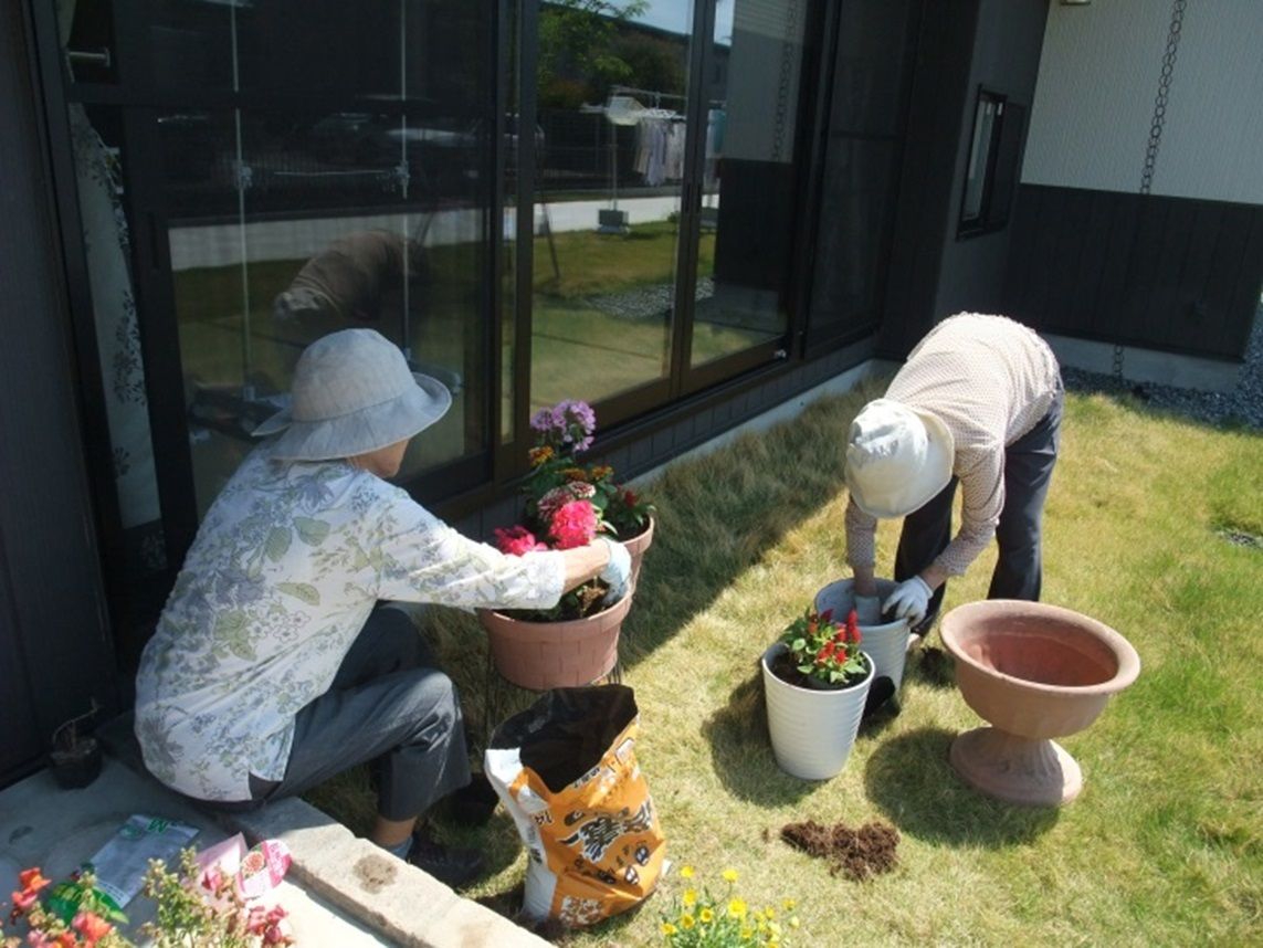 屋外で２人の女性入居者様が、帽子を被り軍手をつけて作業をしている。花の苗を大きな植木鉢に移し替えており、寄せ植えを作っている様子である。