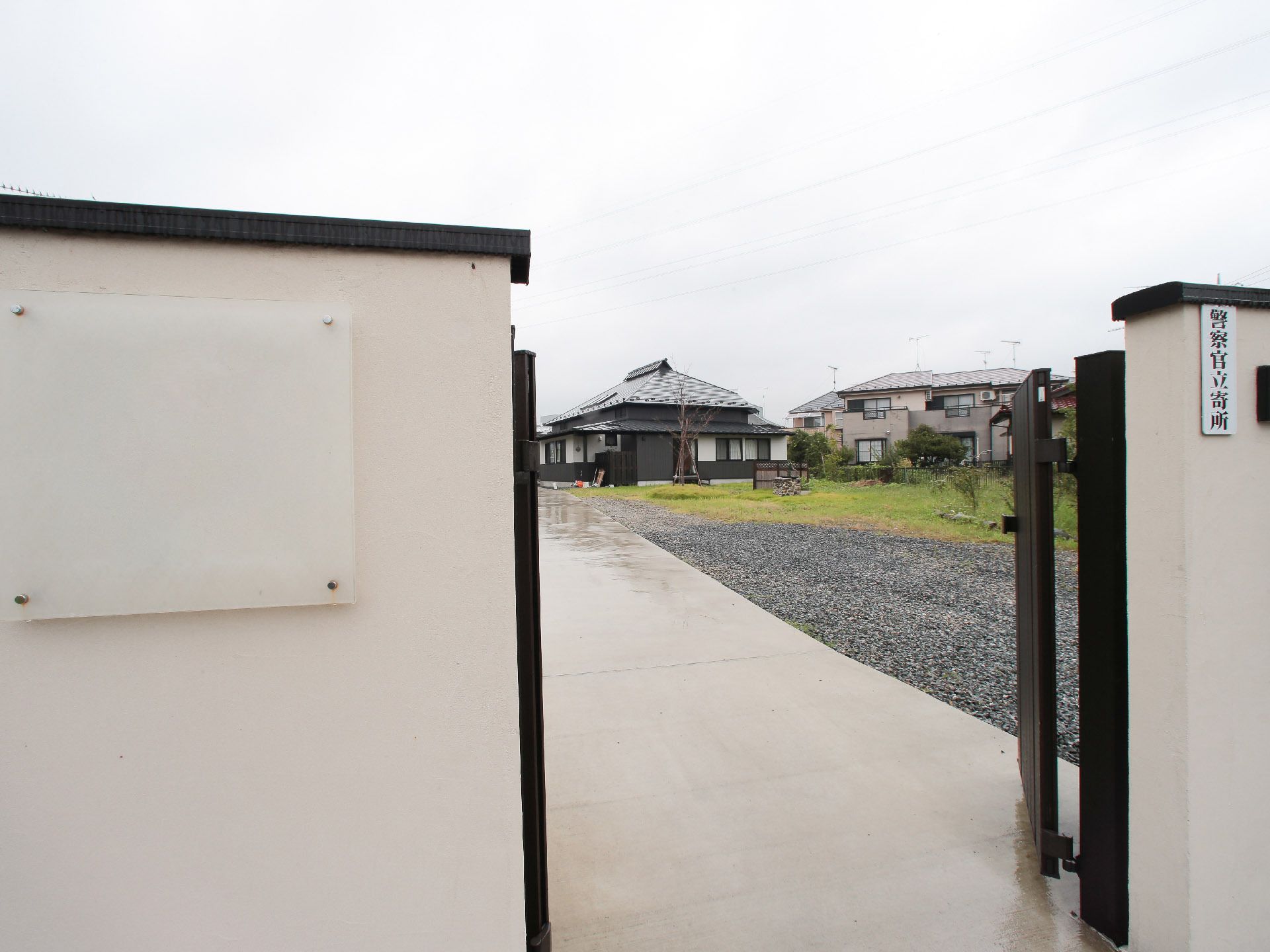 白壁の門構えに表札が掲げてある。白色の背景に黒い墨の字で施設名が書かれている。門には、黒色の格子の扉が取り付けてある。