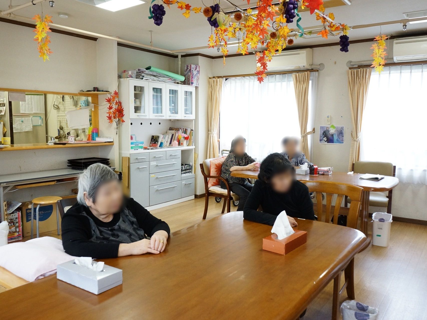 テーブルや椅子が置かれ数名の利用者様が椅子に座っている。天井には製作物が展示されており、秋を感じることができる。