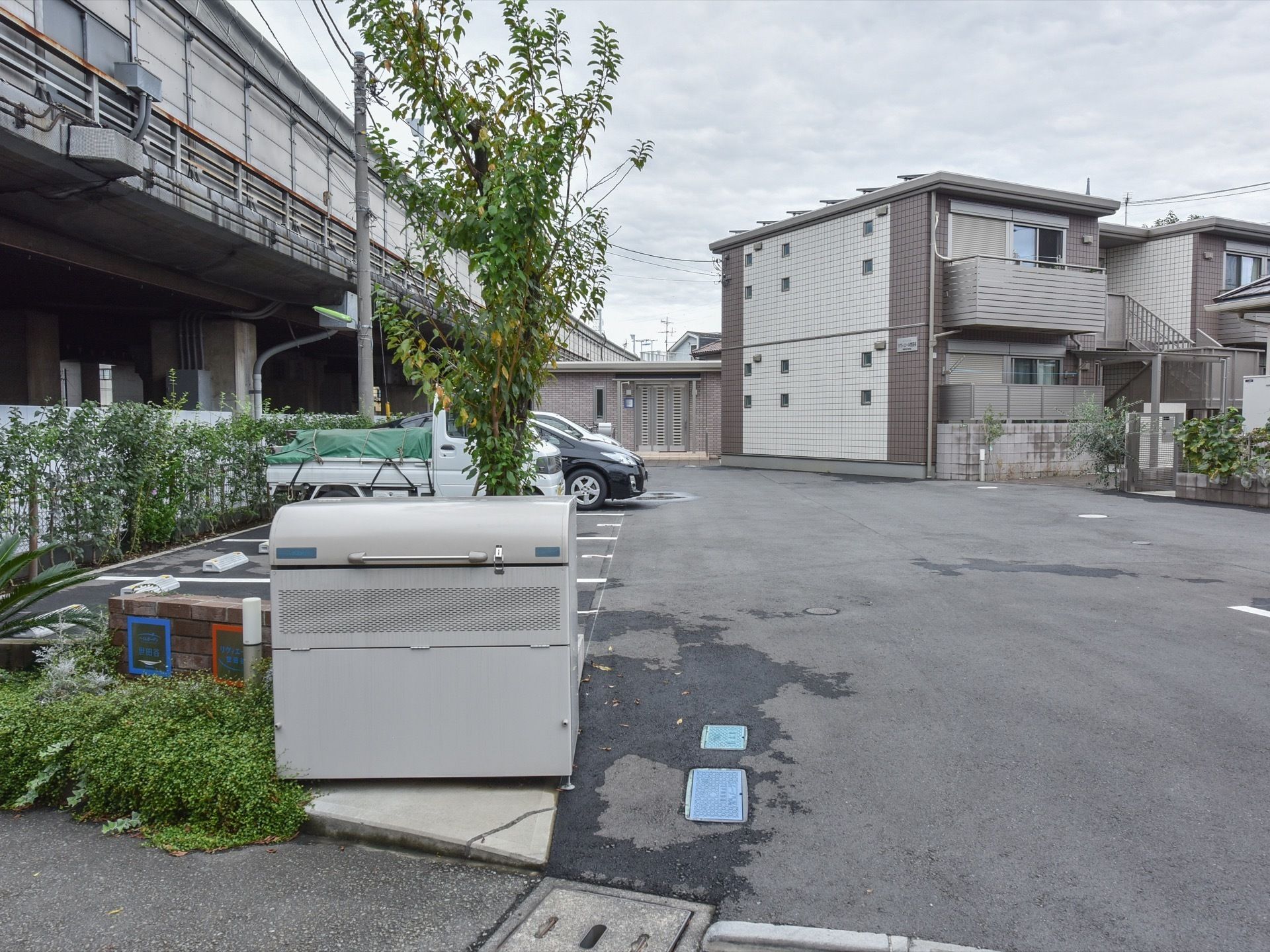 住宅街の中にある２階建ての施設の外観。２階にはベランダが付いており、下まで外階段が設けられている。駐車スペースが見られる。