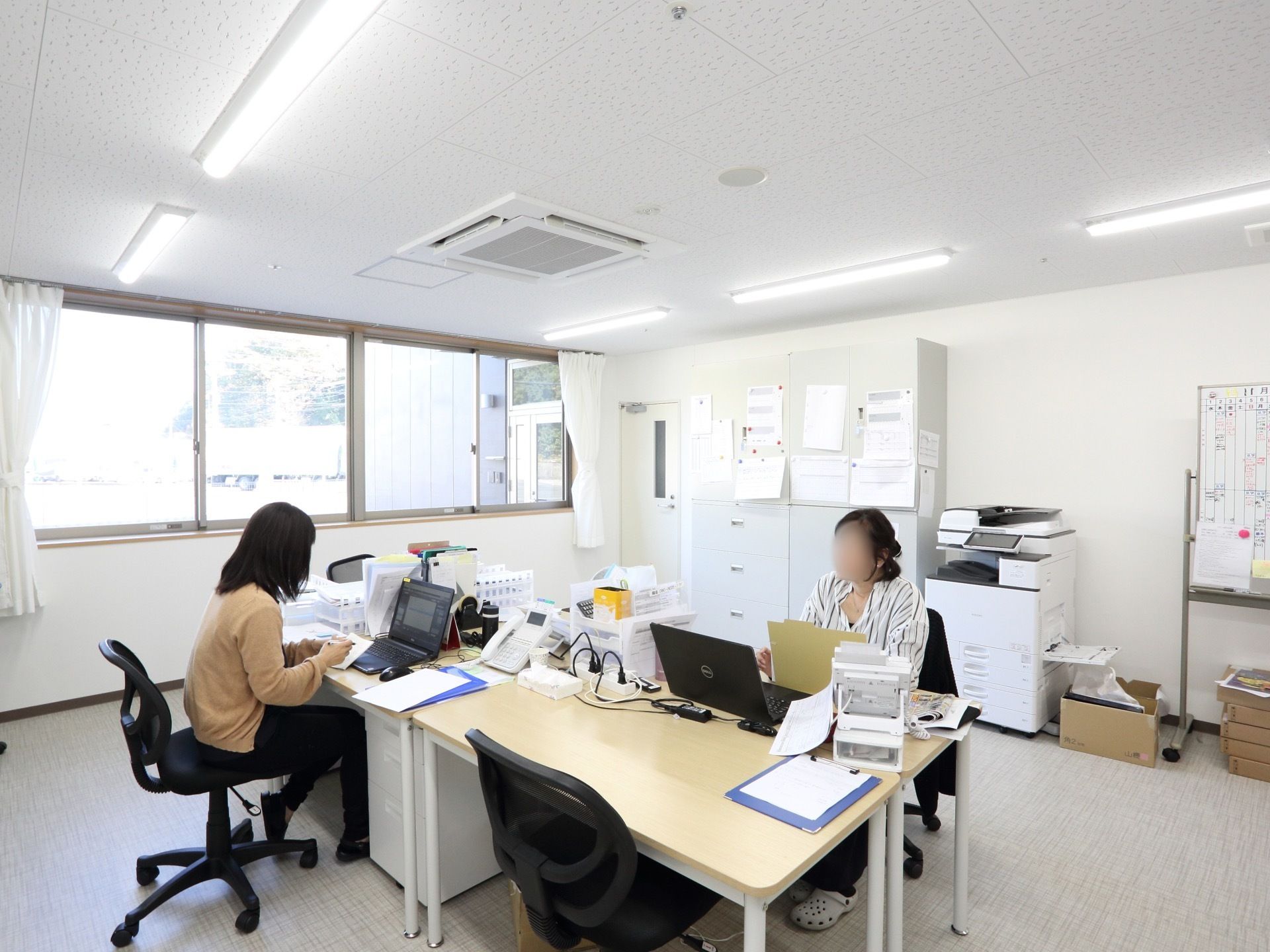 広々とした空間に机と椅子が置かれている。壁側には大きなキャビネット、コピー機、書類が貼られたホワイトボードがある。