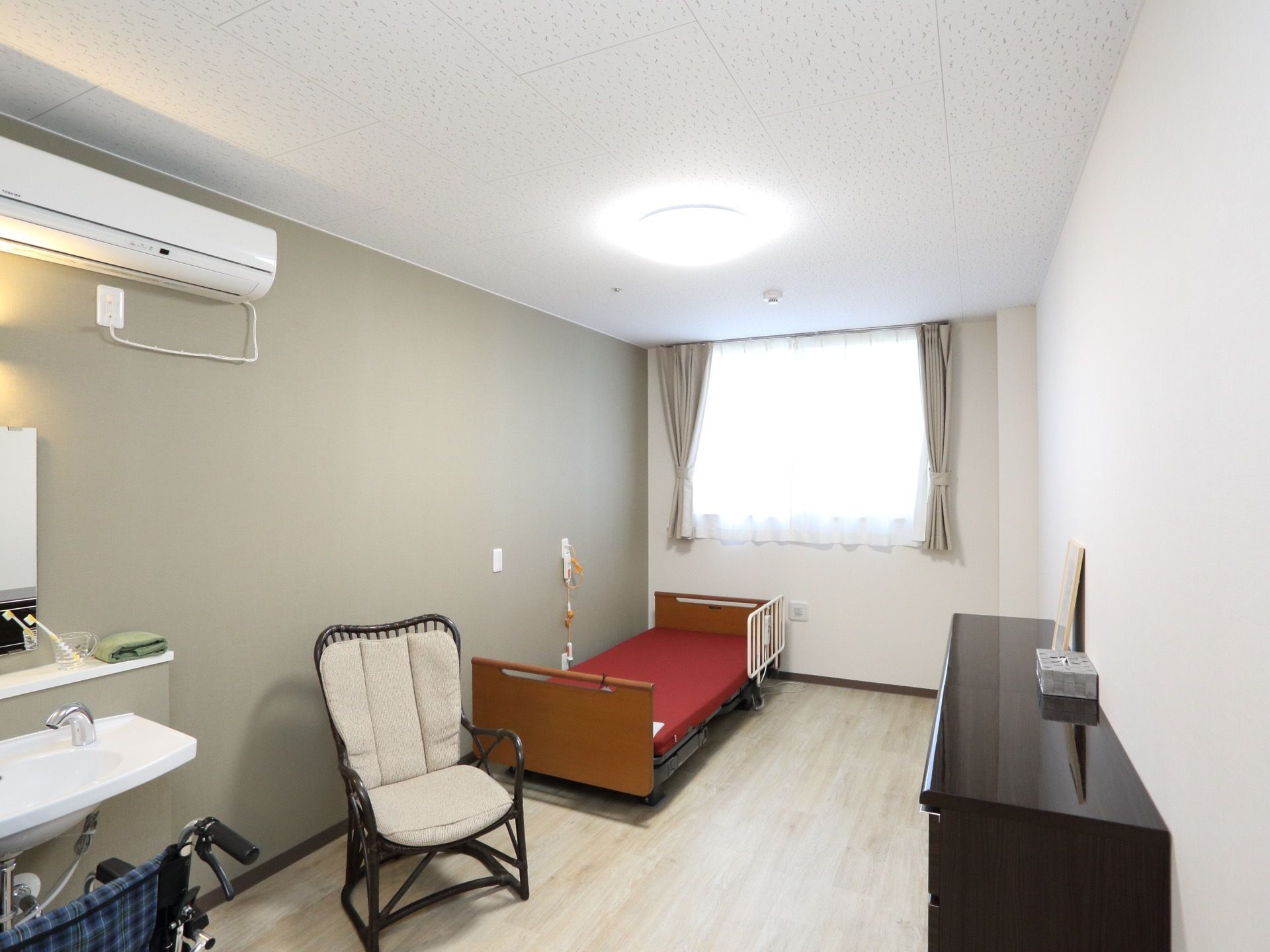 部屋の奥には窓がある。入口側には洗面台とエアコン、窓側には介護用のベッドが置かれている。壁にはナースコールがある。