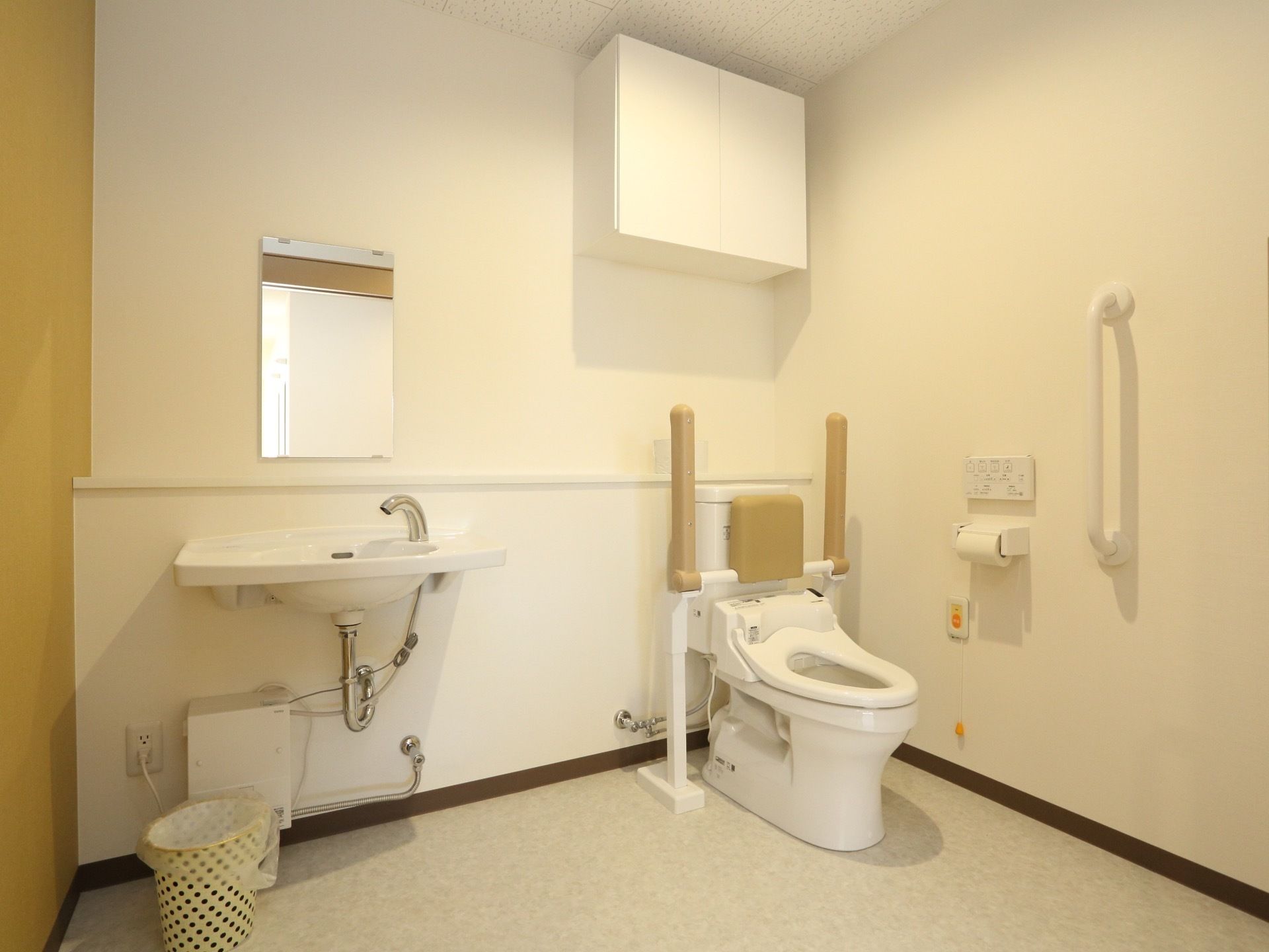 広い空間にトイレと洗面台が配置されている。壁にはナースコールや手すりがあり、上には収納棚が設置されている。