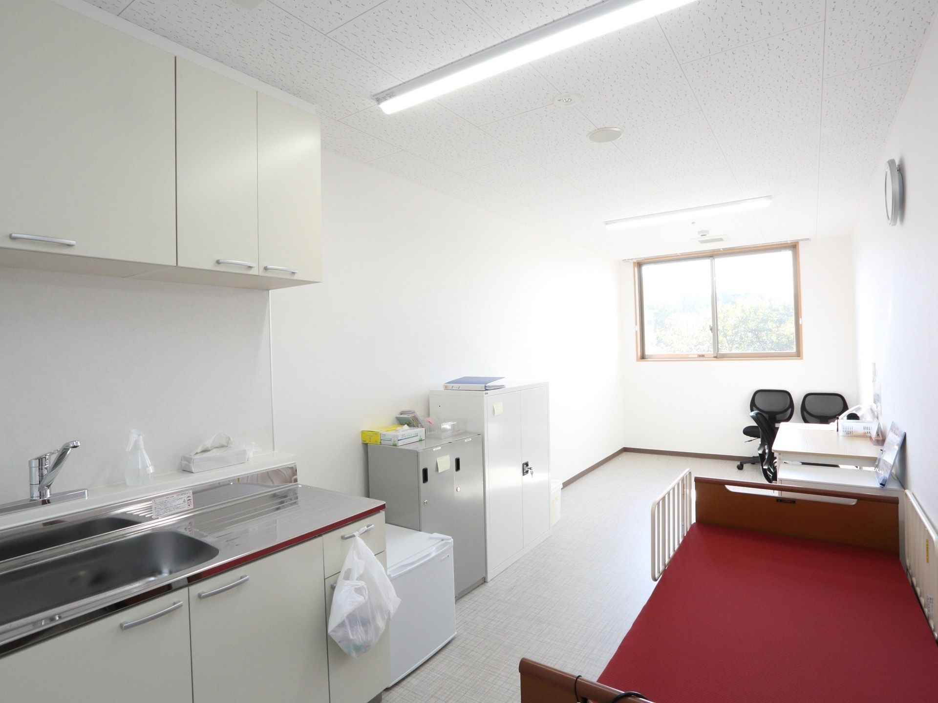 奥行きのある部屋にベッドと冷蔵庫、机と椅子が置かれている。ベッドの近くにはシンクがある。ファイルなどをしまう棚もある。