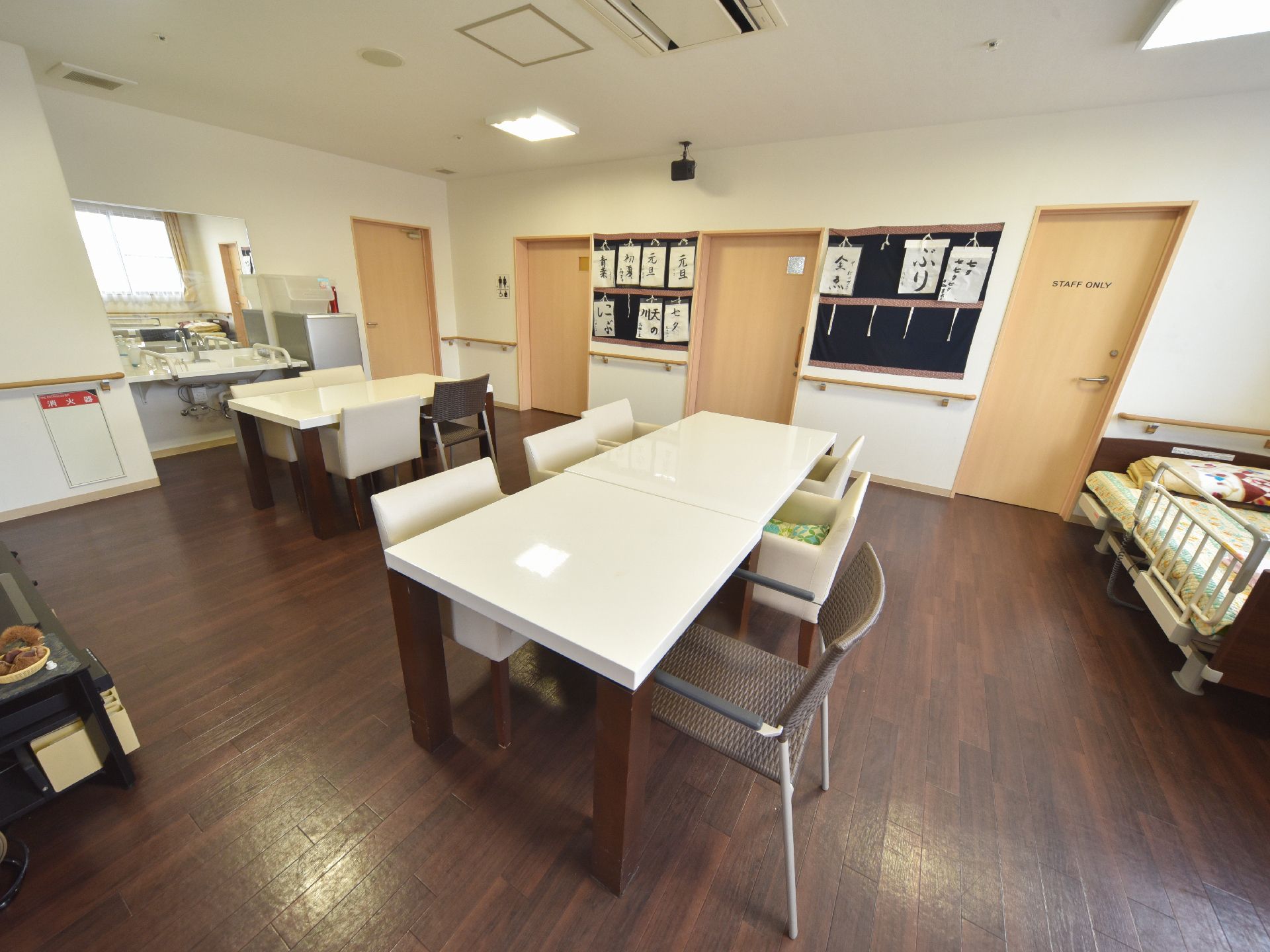 テーブルと椅子が複数置かれている。床は濃い茶色のフローリングになっている。壁や天井には白色が塗られている。