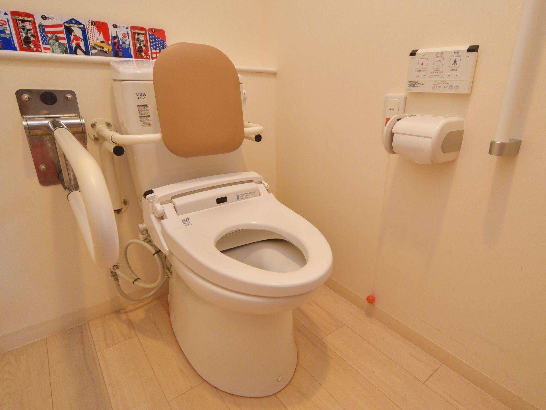 背もたれ付きの便器が正面にある。左側には手すりが付いている。右側の壁には温水便座洗浄機の操作パネルがある。