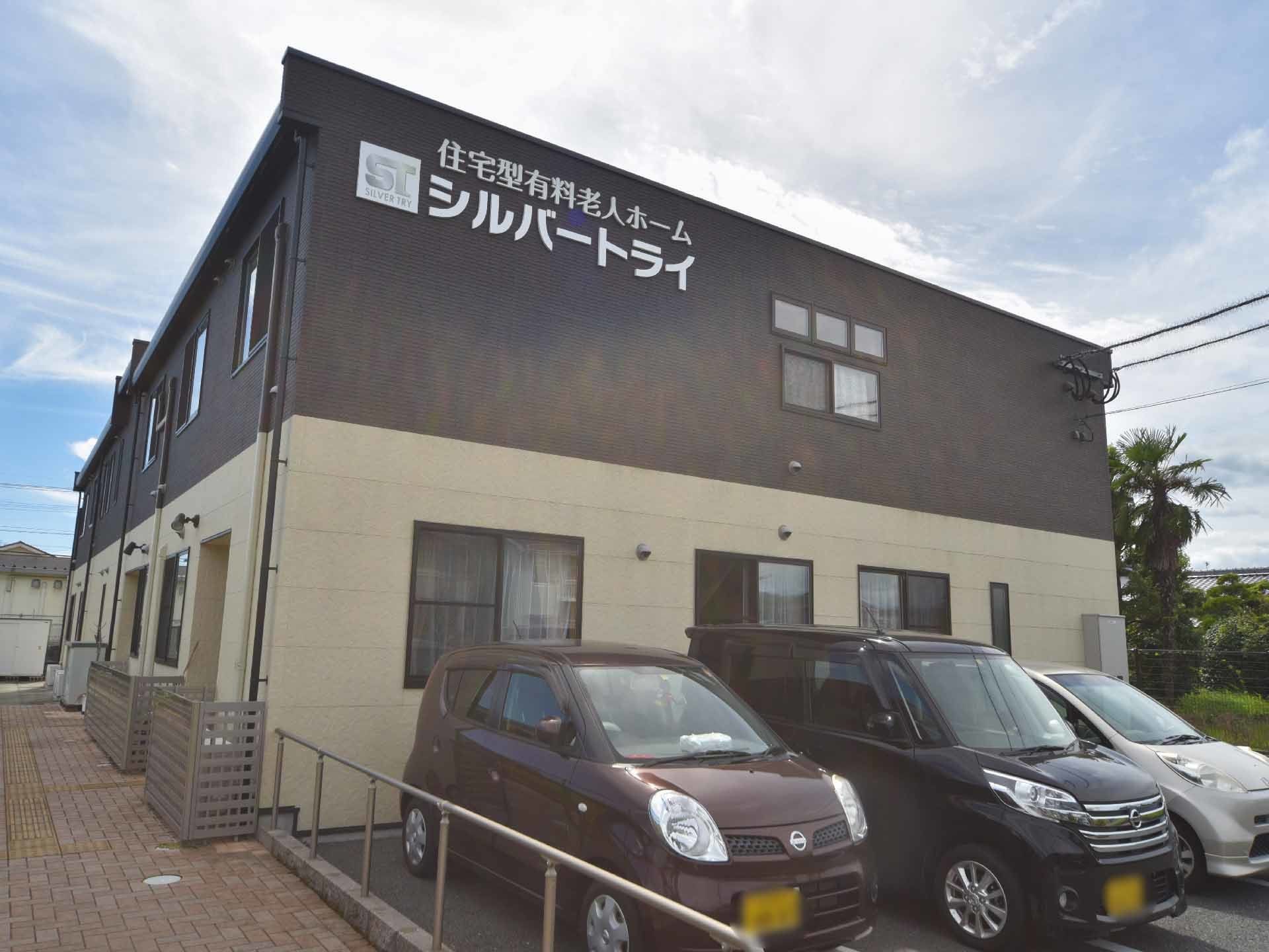 正面に建物が建っており、その手前には駐車場がある。外壁は黒色とクリーム色の2色で塗られている。そこの左上側には施設名が書かれている。