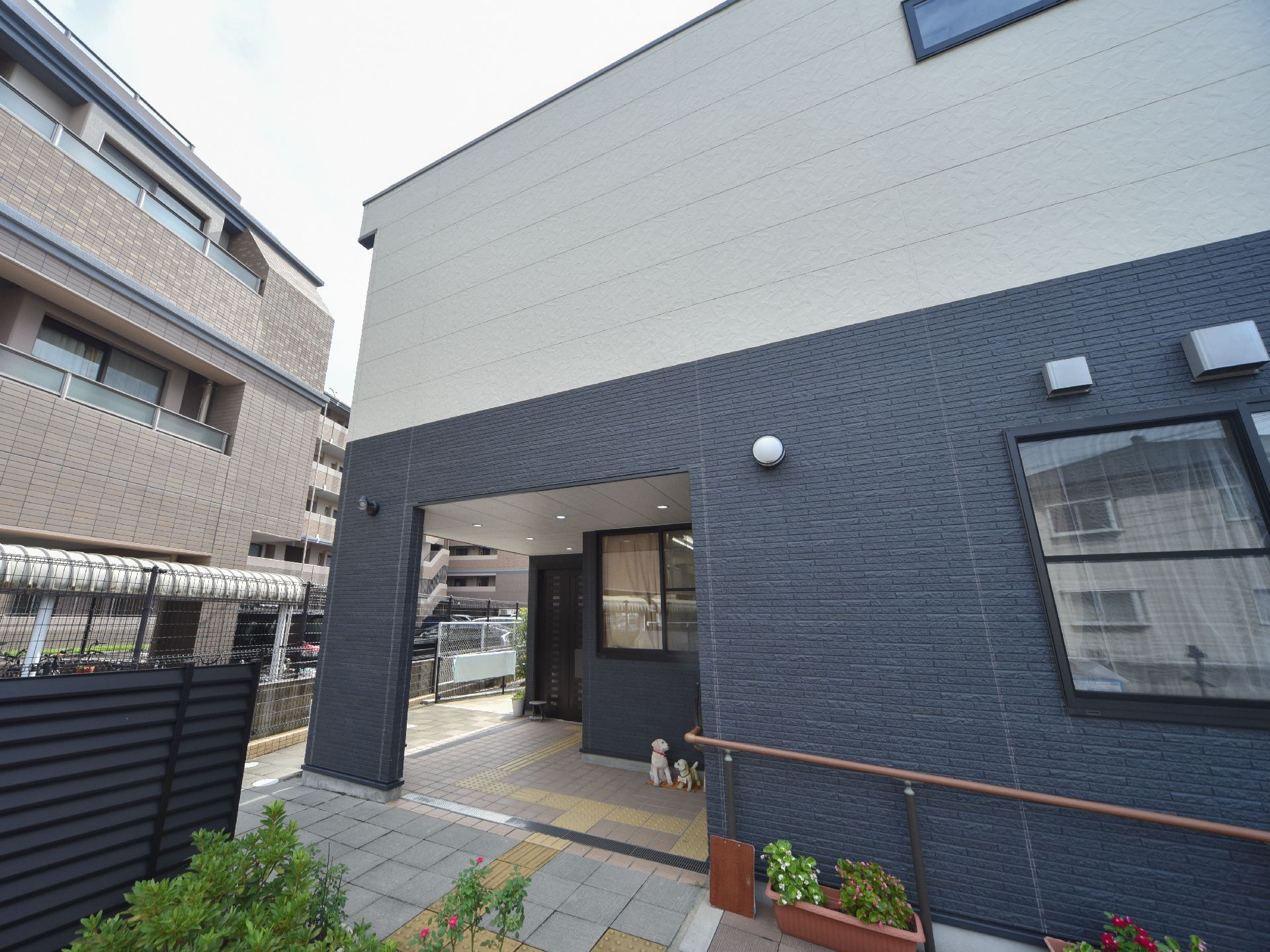 正面に建物がある。右側から外壁に沿って手すりが付いており、その下側には植物が植えられたプランターが置かれている。