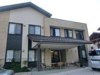 二階建てのベージュ系の外観の建物で、玄関前には瓦屋根風のひさしが付いている。ひさしの下はテラスになっており、テーブルや椅子が置かれている。