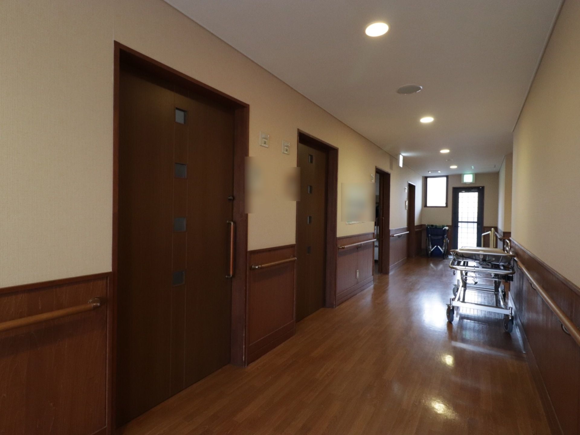 フローリングや居室のドアは濃い茶色で統一されている。廊下の奥に車椅子が置いてある。ストレッチャーのようなものが置いてある。