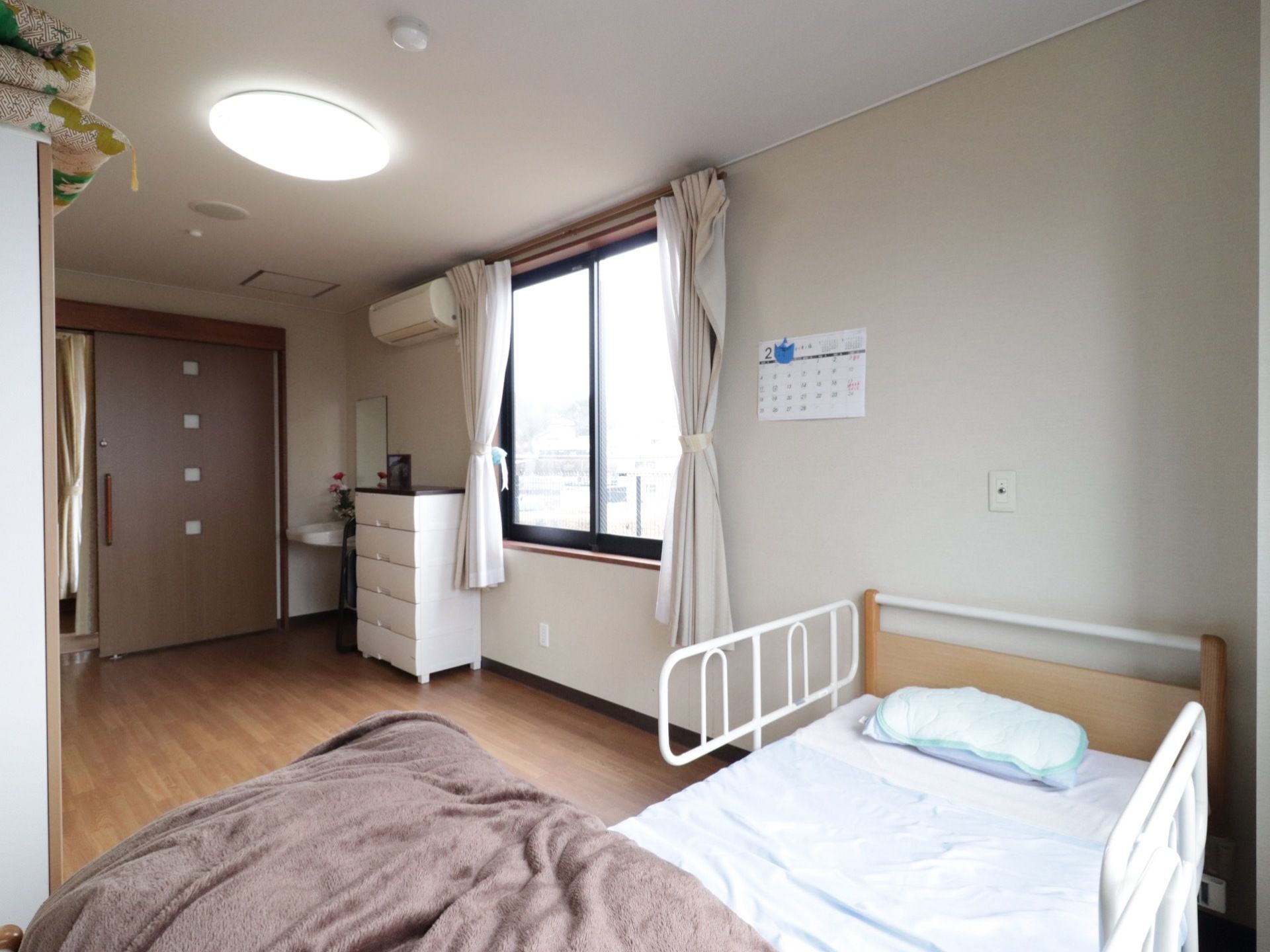 入口の横に洗面台が設置してあり、その横には5段の衣装ケースが置いてある。ベッドが１台置いてある。たんすのようなものがある。