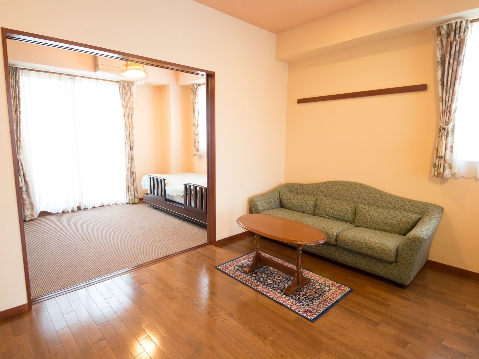奥の部屋にはベッドがあり、手前の部屋には３人がけのソファとテーブルが置かれている。窓には、花柄のカーテンがつけられている。