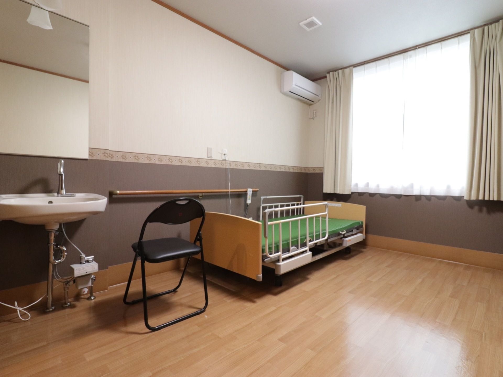 部屋の奥には高さのある窓がある。壁側には、手すりと介護用のベッド、パイプ椅子が置かれている。ナースコールもある。