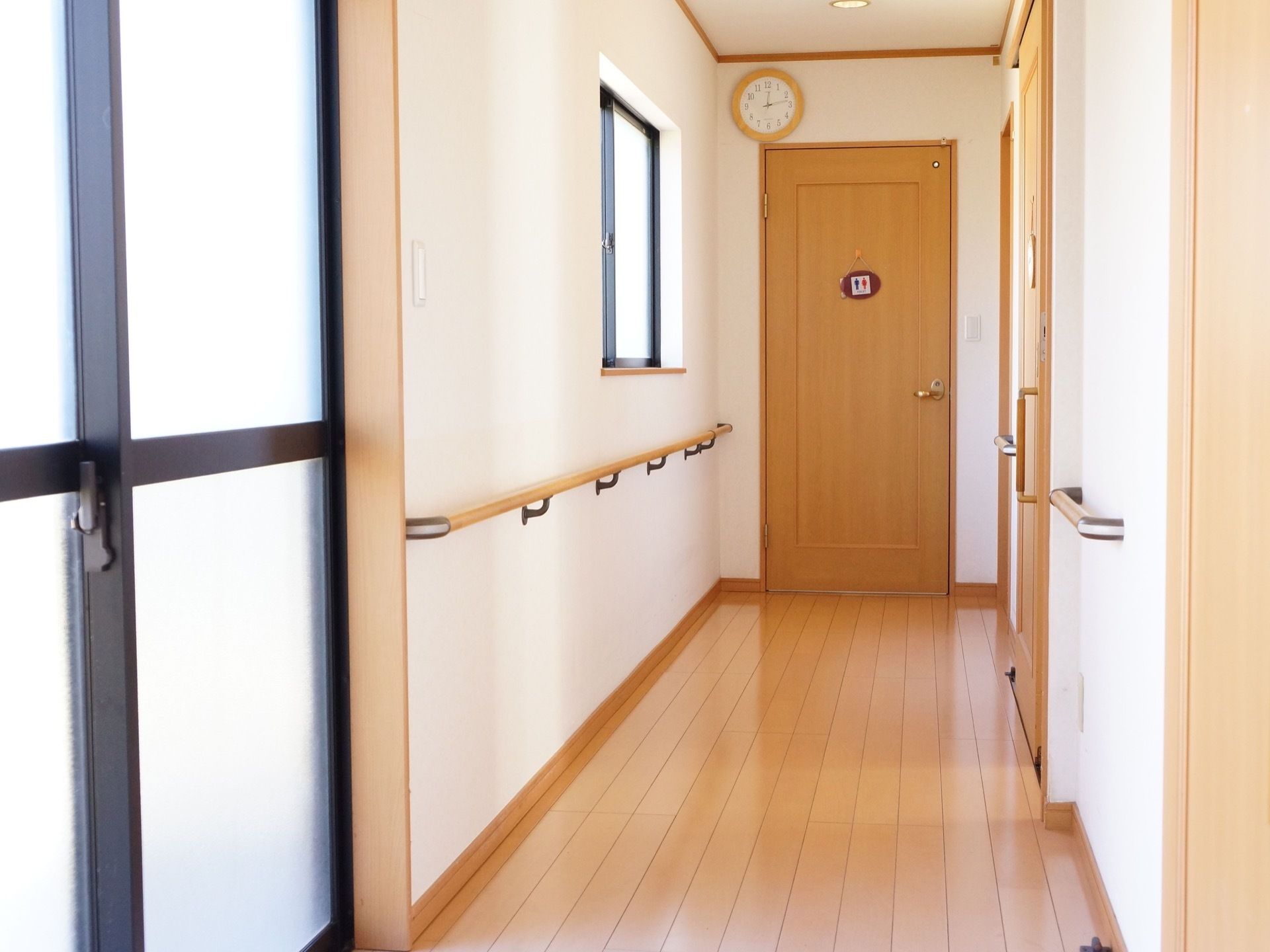 手すりが取り付けられた廊下の突き当たりに、トイレのサインがかかったドアがある。ドアの上には時計をかけている。