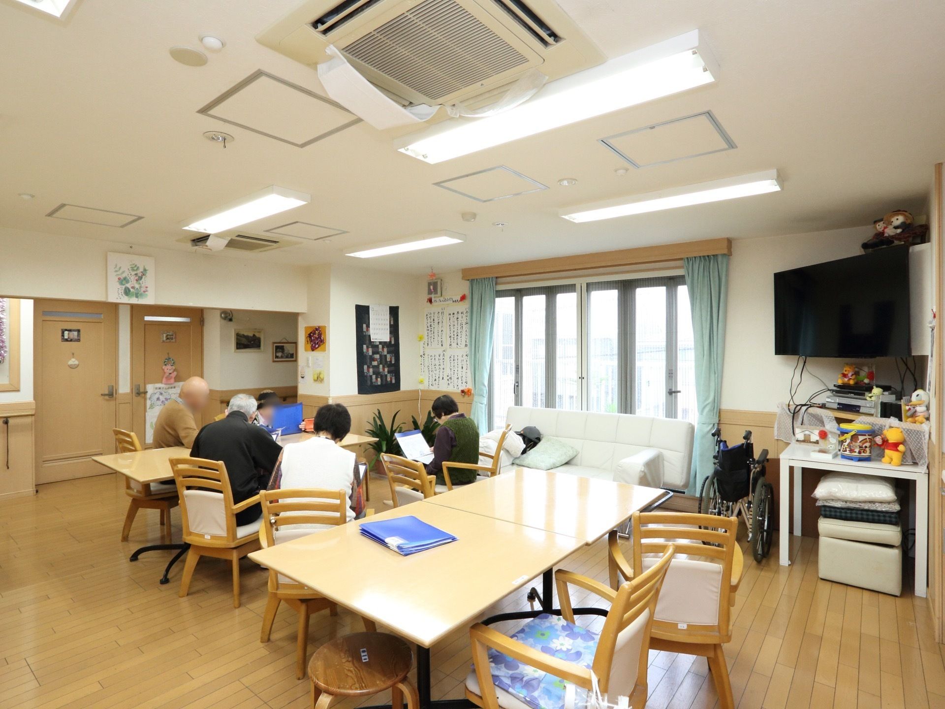 テーブルと椅子が置かれており、利用者様が集まっている。ソファ、大型テレビが配置されている。ドアのないオープンスペースである。