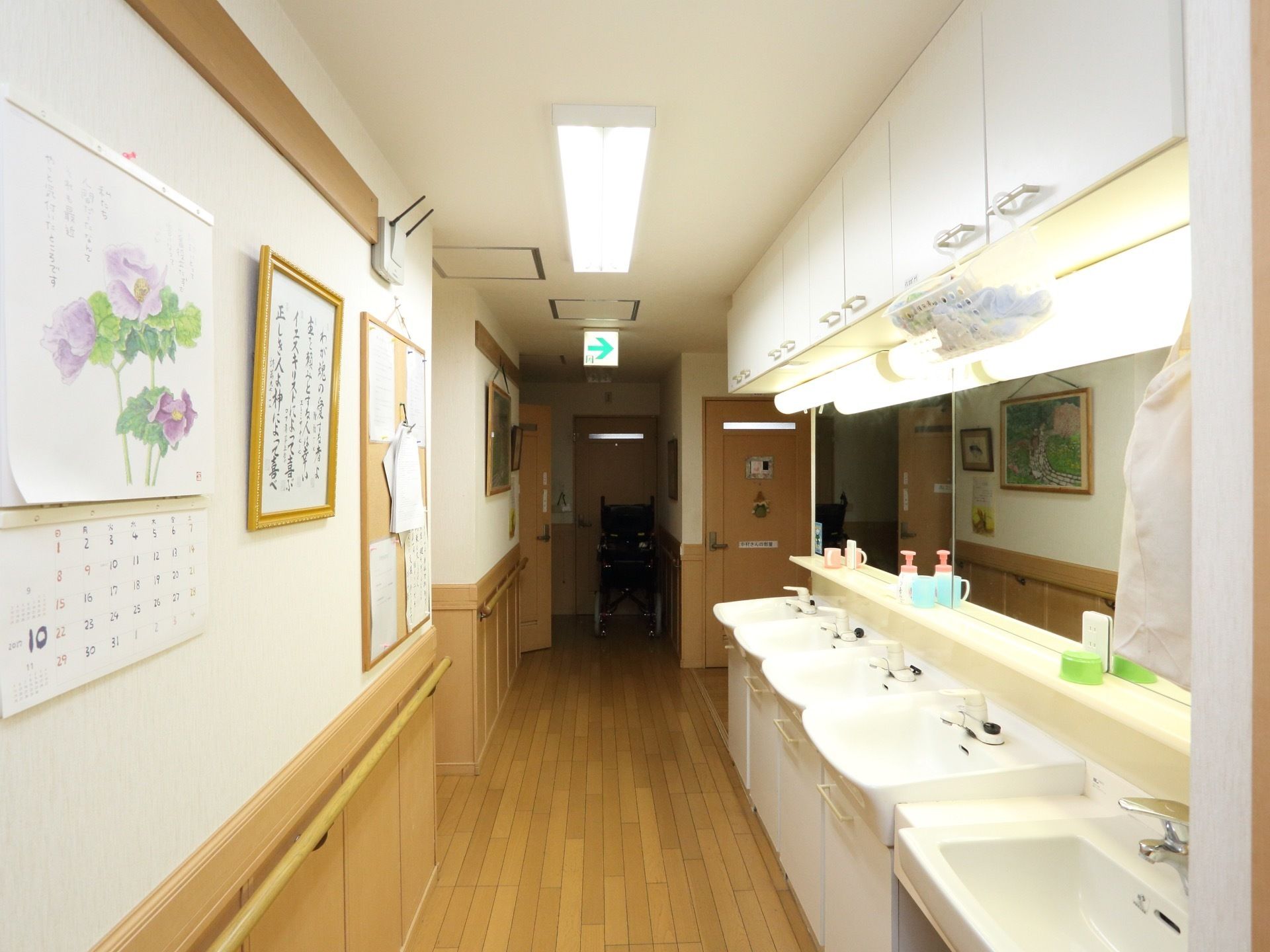 壁に手すりが設置してある。絵画やカレンダーが掛けられている。５台の鏡付き洗面台がある。非常口の表示がある。